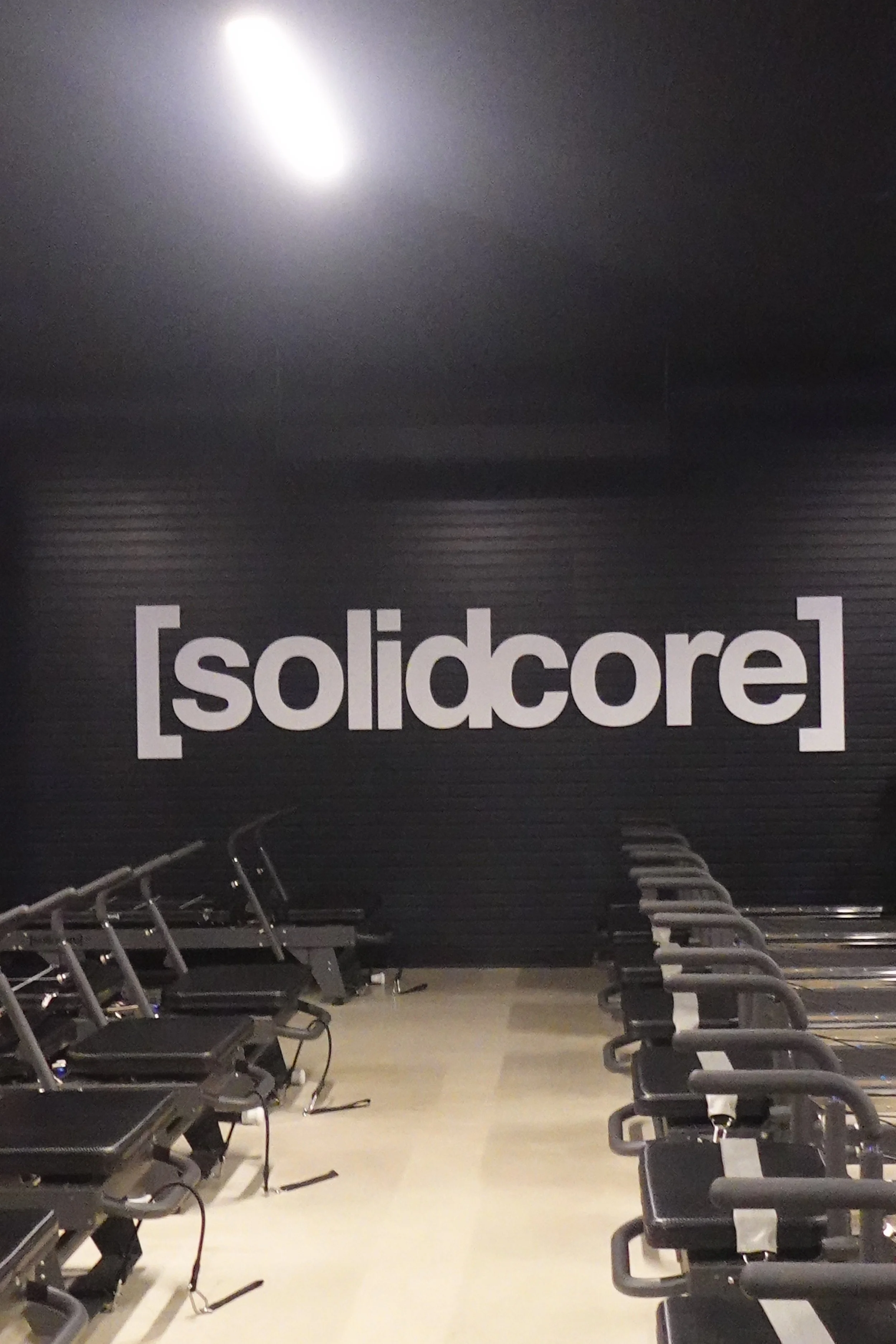 Empty gym with black wall featuring the [solidcore] logo in large letters and multiple exercise machines lined up on both sides.