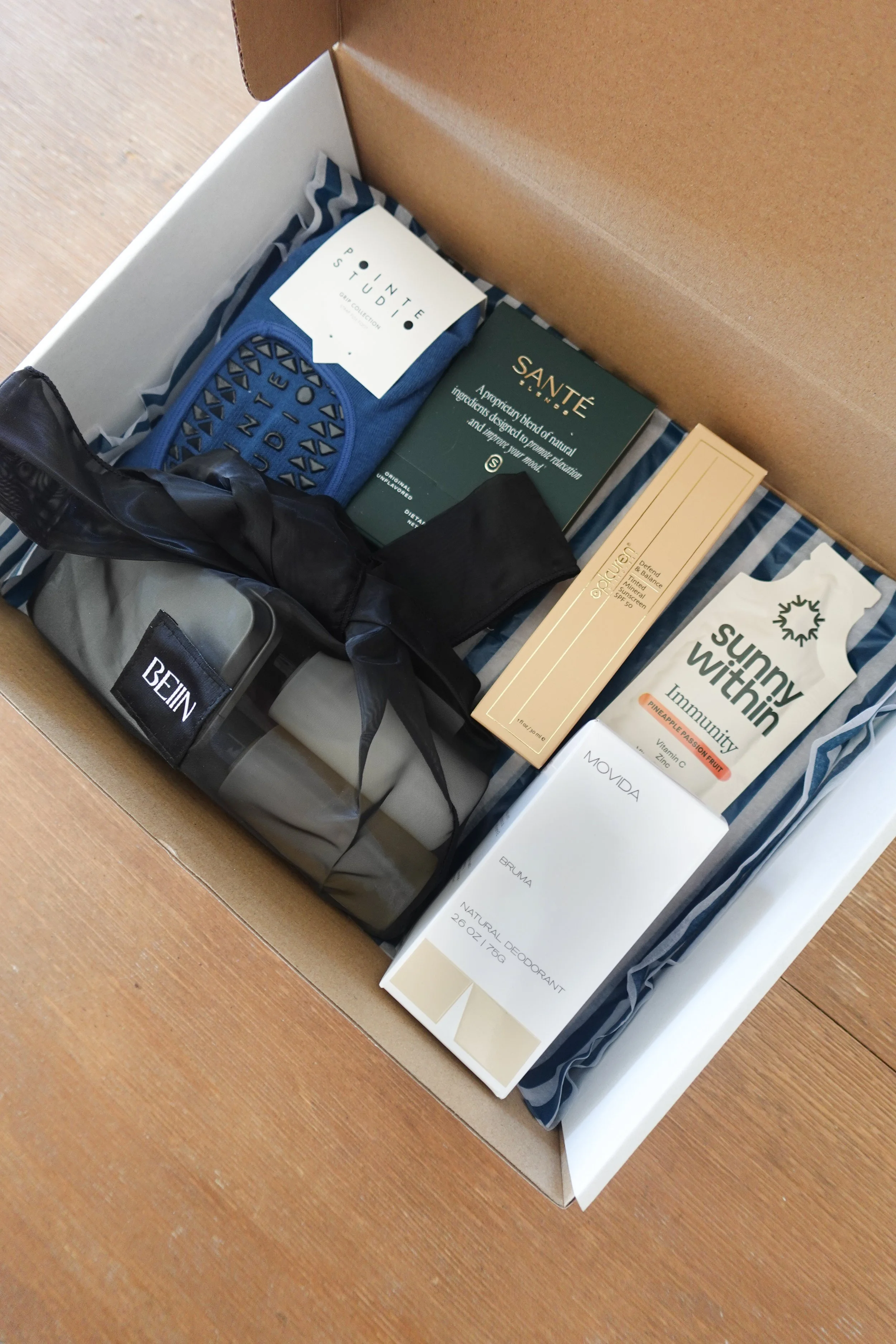 Open gift box containing various personal care and wellness items, including a bottle of sunscreen, natural deodorant, immunity supplement, skincare products, a pouch, and a small bag, all arranged on a wooden surface.
