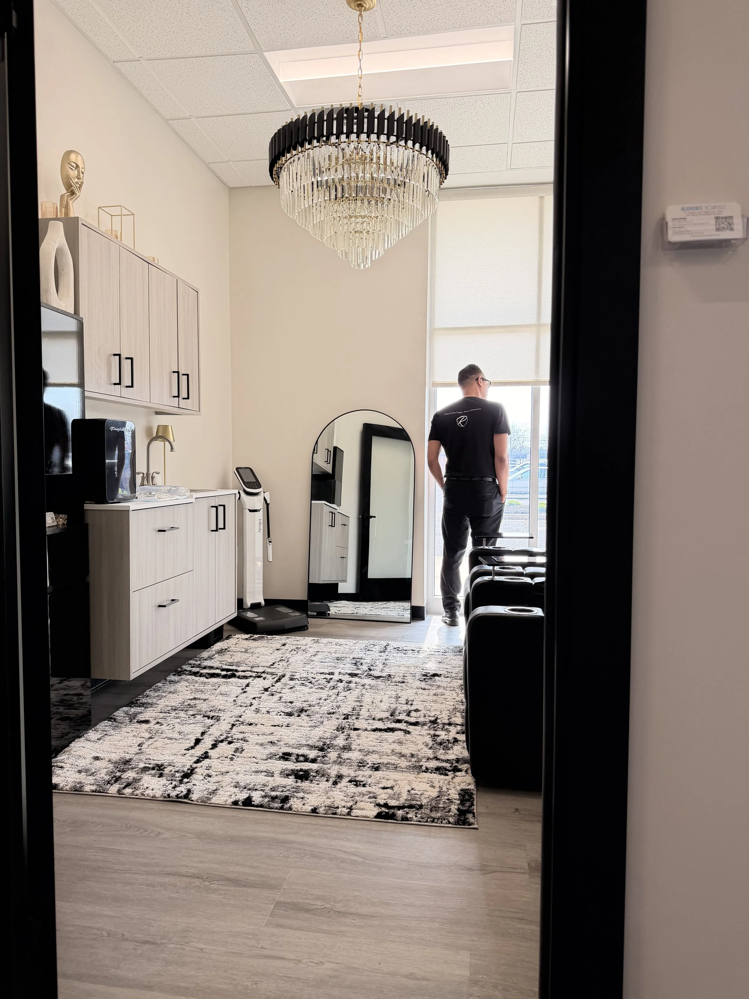 Office space with a chandelier, a man in black shirt and grey pants looking out a large window, a mirrored full-length mirror, a cabinet with decor, a small fridge, a cordless phone, and a patterned rug on light wood flooring.