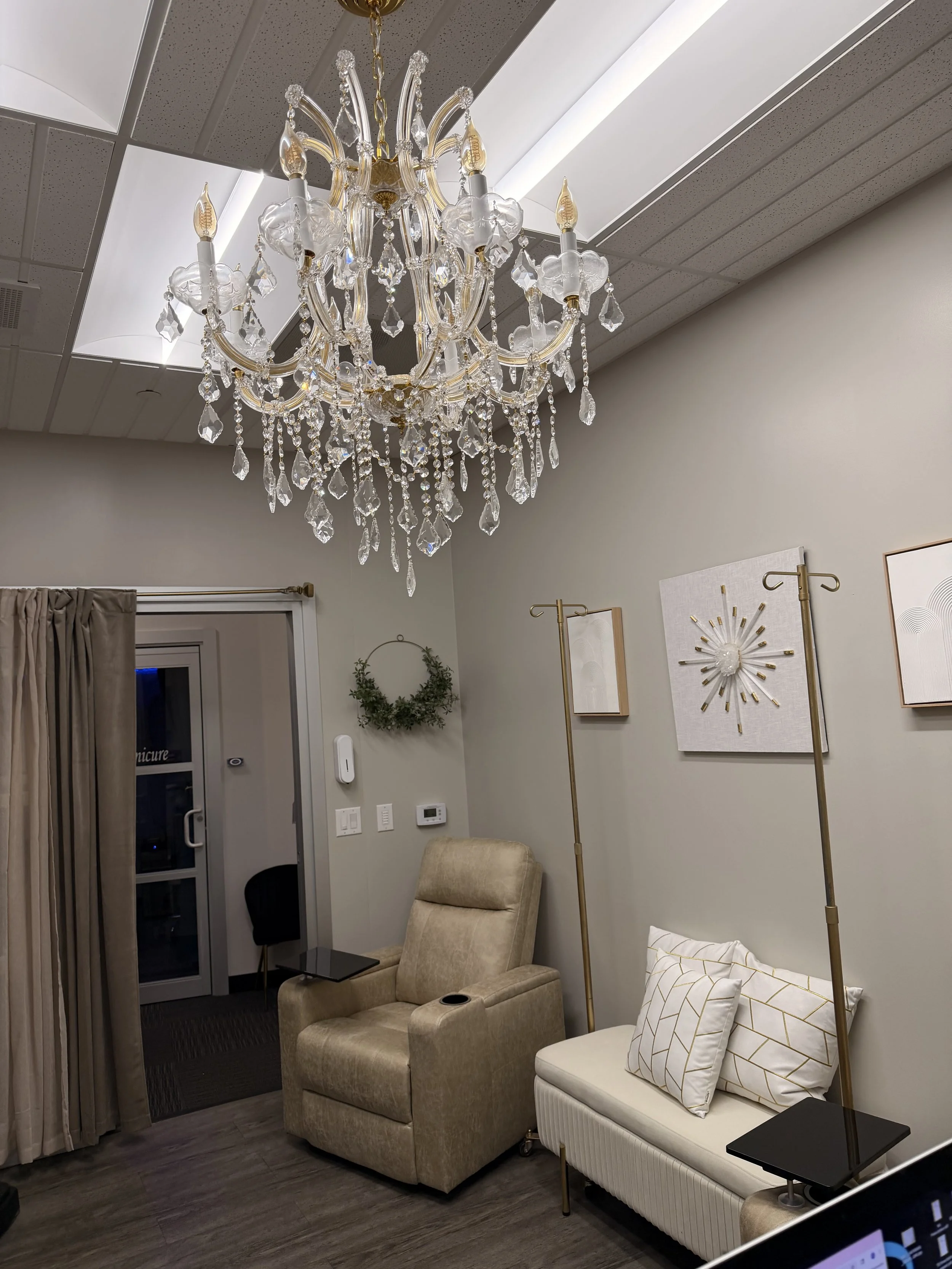 Interior of a room with a large crystal chandelier hanging from the ceiling, cream-colored recliner, white bench with pillows, and decorative wall art with gold accents.
