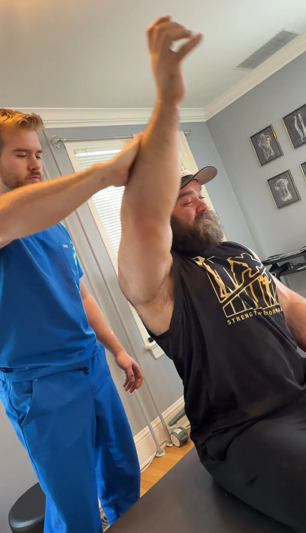 A man with a beard and a cap performs a stretching exercise with his arm raised while a healthcare professional in blue scrubs assists him in a room with framed pictures of skeletons on the wall.