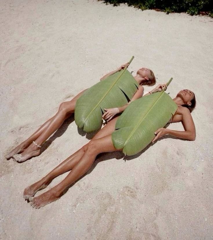 Two women lying on the beach covered with large green leaves, holding drinks, and relaxing.