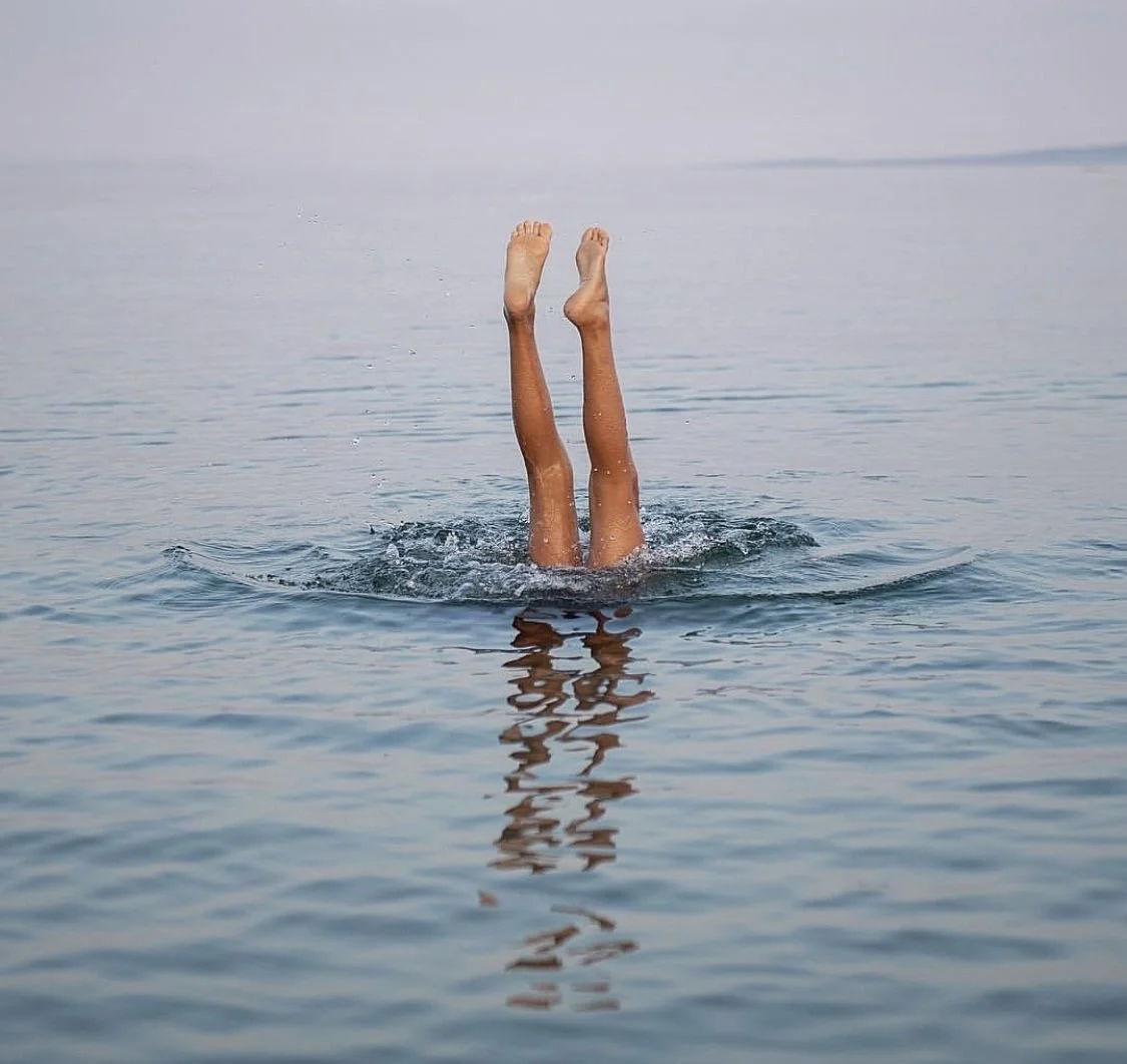 A person submerged in water with only their legs and feet visible, raised vertically above the water surface, creating a reflection in the water.