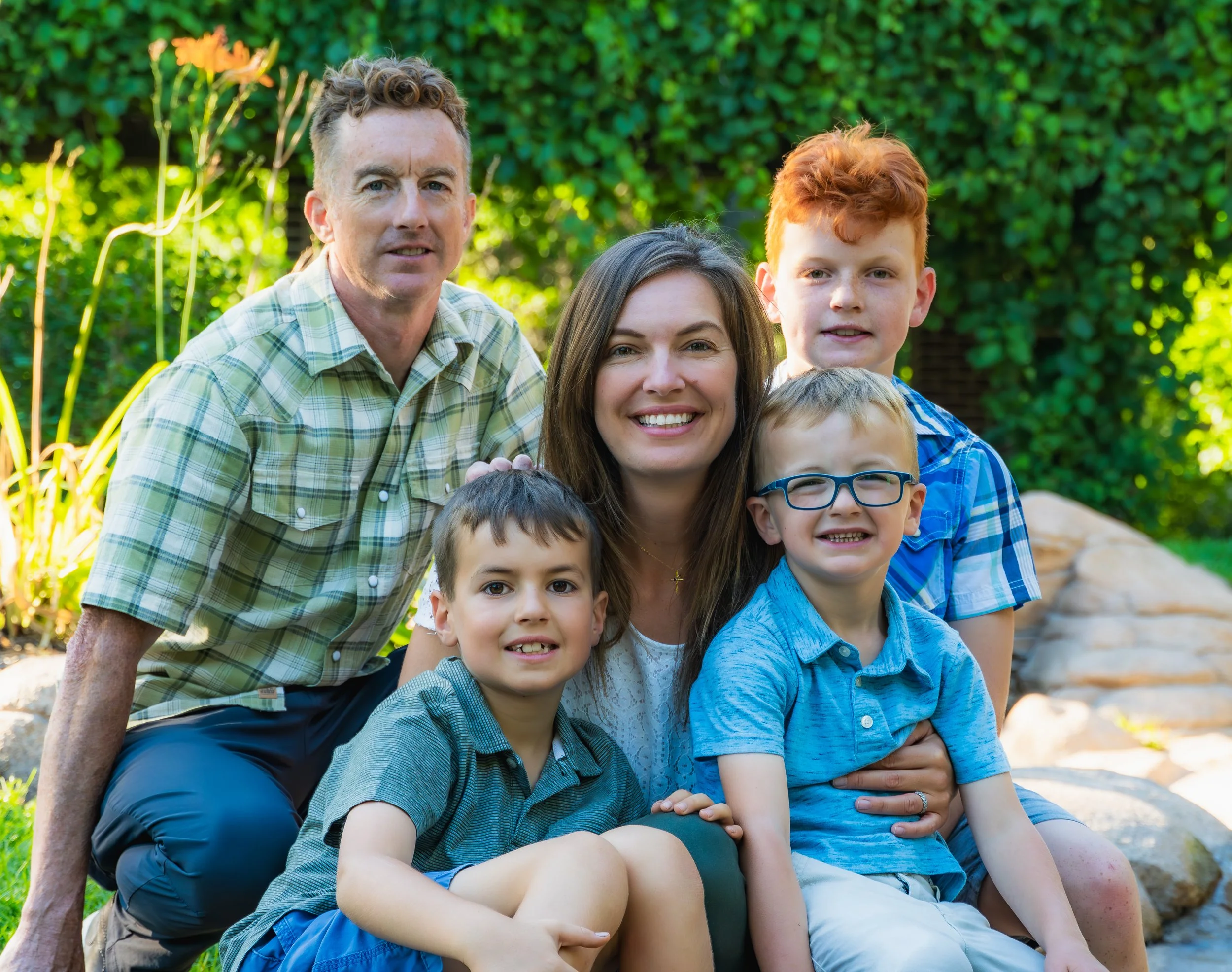 A family portrait of five posing outdoors in a garden, smiling and sitting on rocks with greenery and flowers in Minneapolis park.