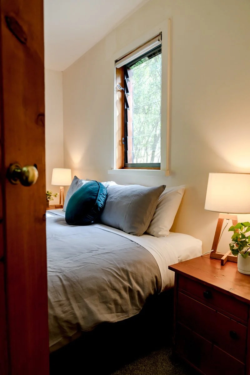 Cozy bedroom with bed, gray bedding, teal pillow, wooden nightstand, lamp, and window view.