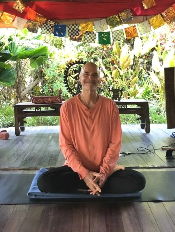 Steve Ross meditating in a peaceful, rustic setting in Bali with prayer flags and tropical plants