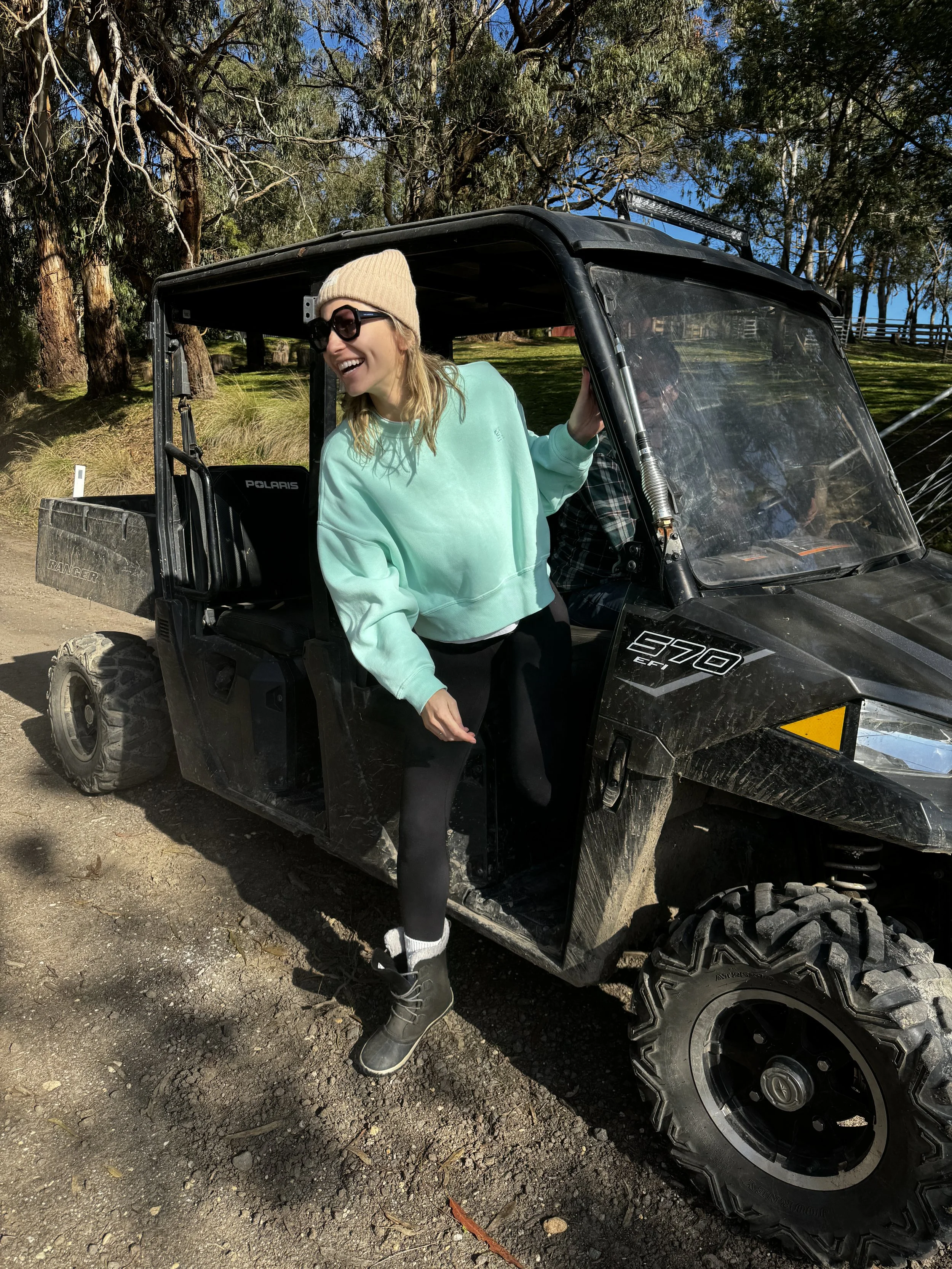 Person in a mint sweatshirt, black leggings, winter boots, and a beige beanie smiling while stepping out of an off-road vehicle on a dirt road.
