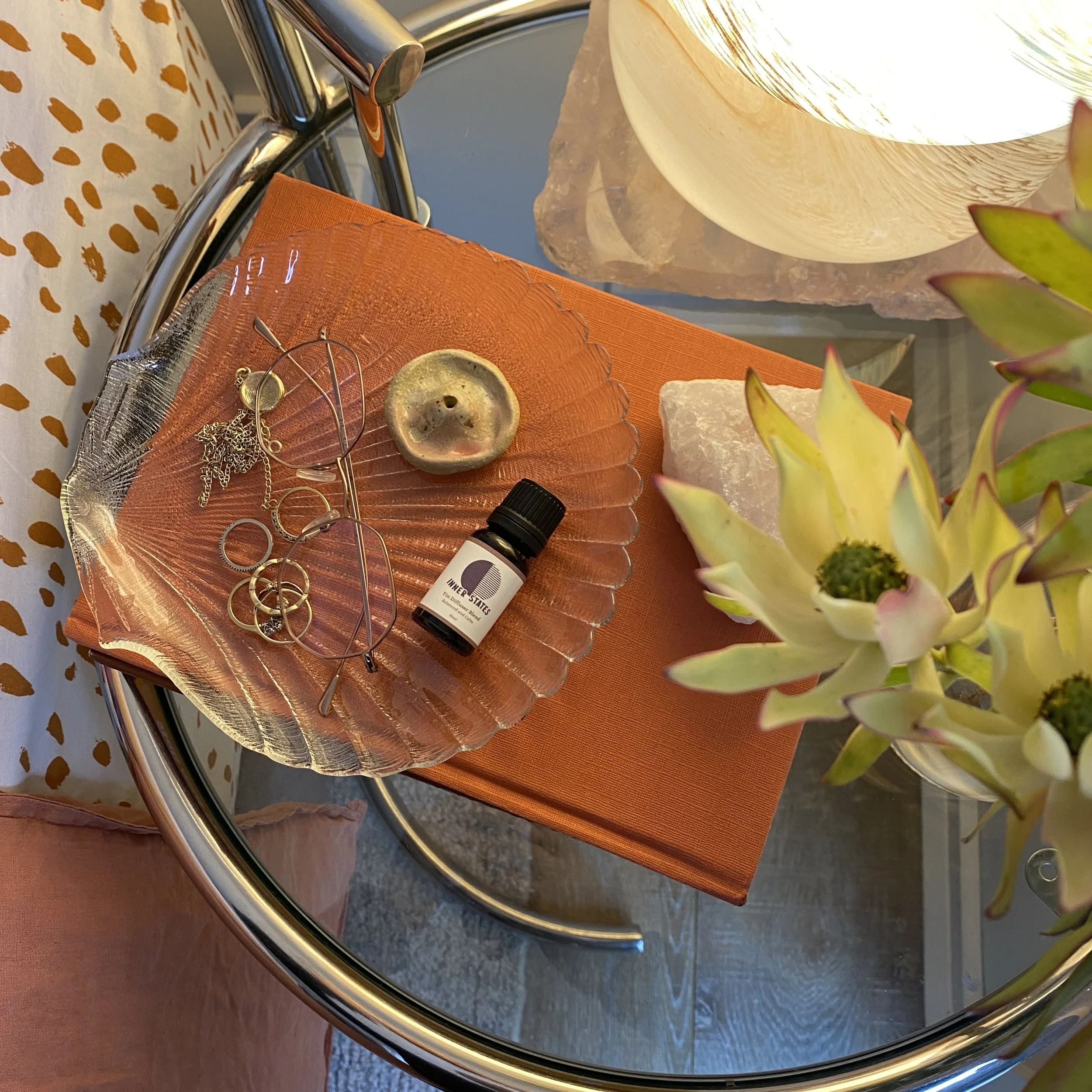 Decorative table setup with a glass dish holding jewelry, eyeglasses, an essential oil bottle labeled "Antique Thief," surrounded by a decorative notebook and flowers.