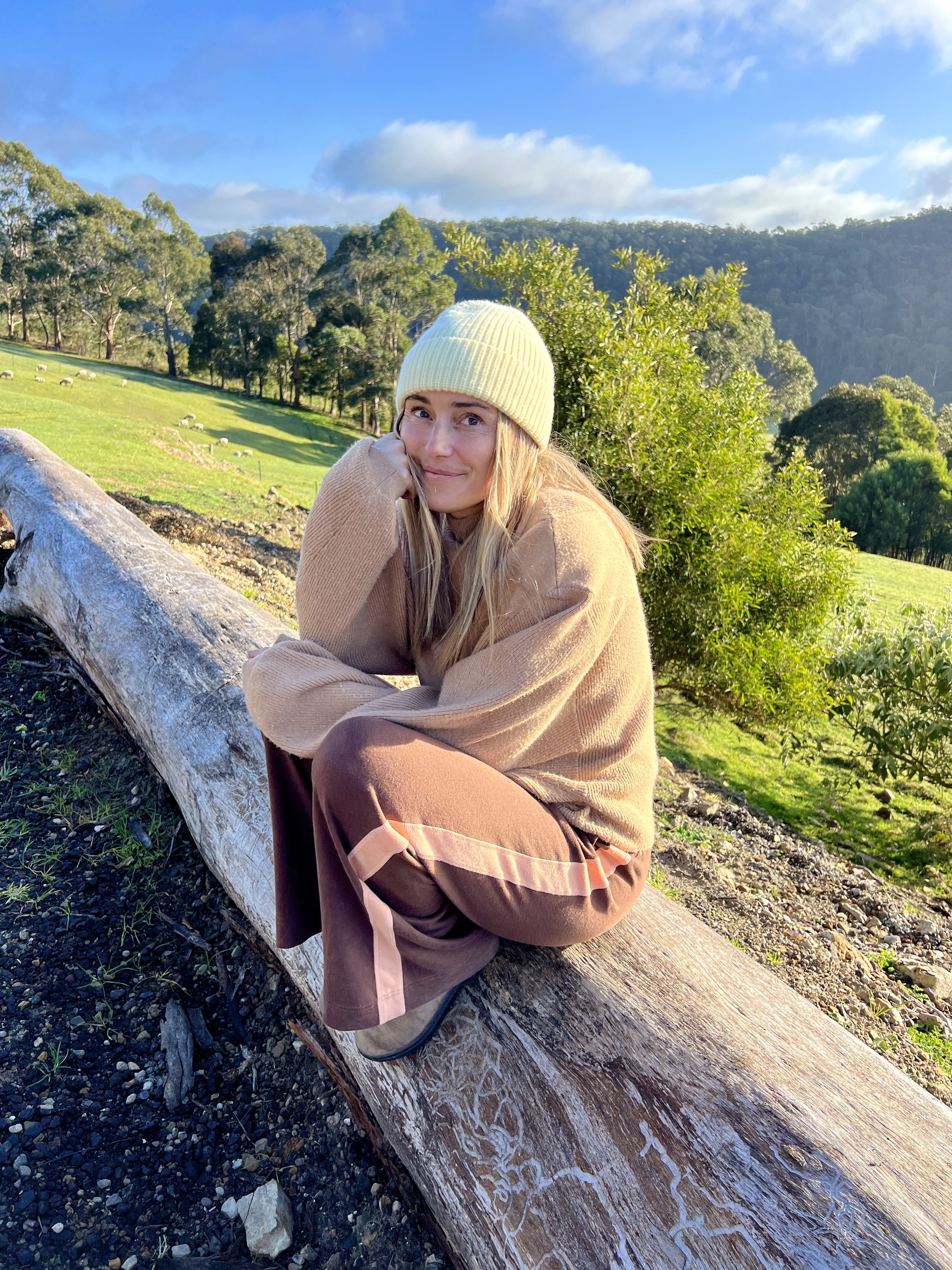 Lauren Williams of Will and Bear  in a beanie sitting on a log, with green landscape and trees in the background.