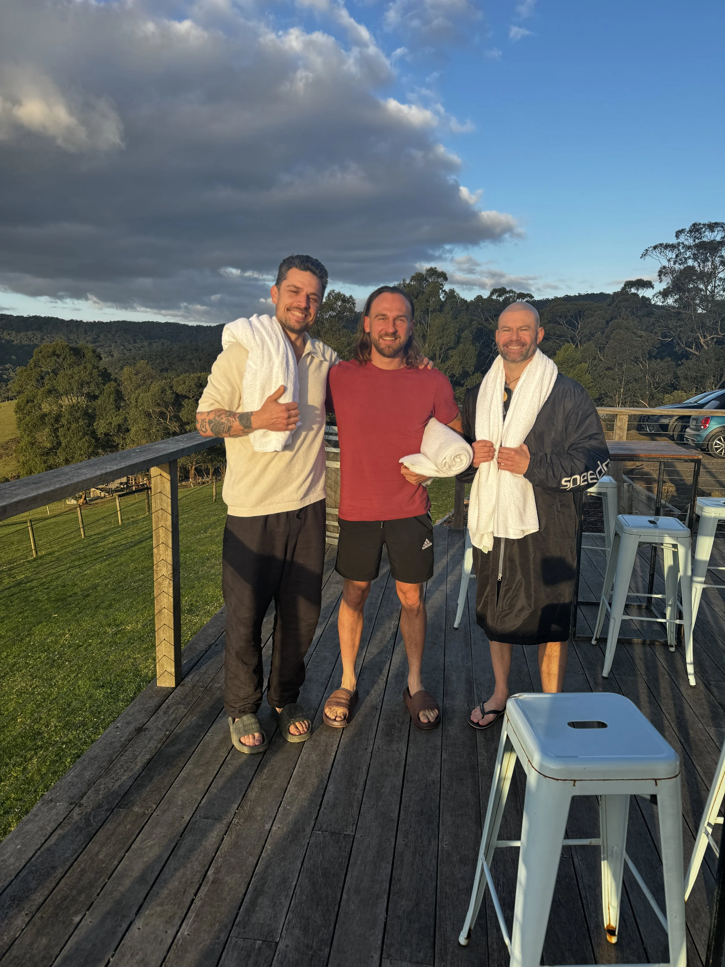 Three men standing on a wooden deck with towels around them, outdoor setting with trees and a cloudy sky in the background. This is King Parrot cottages in The Otways