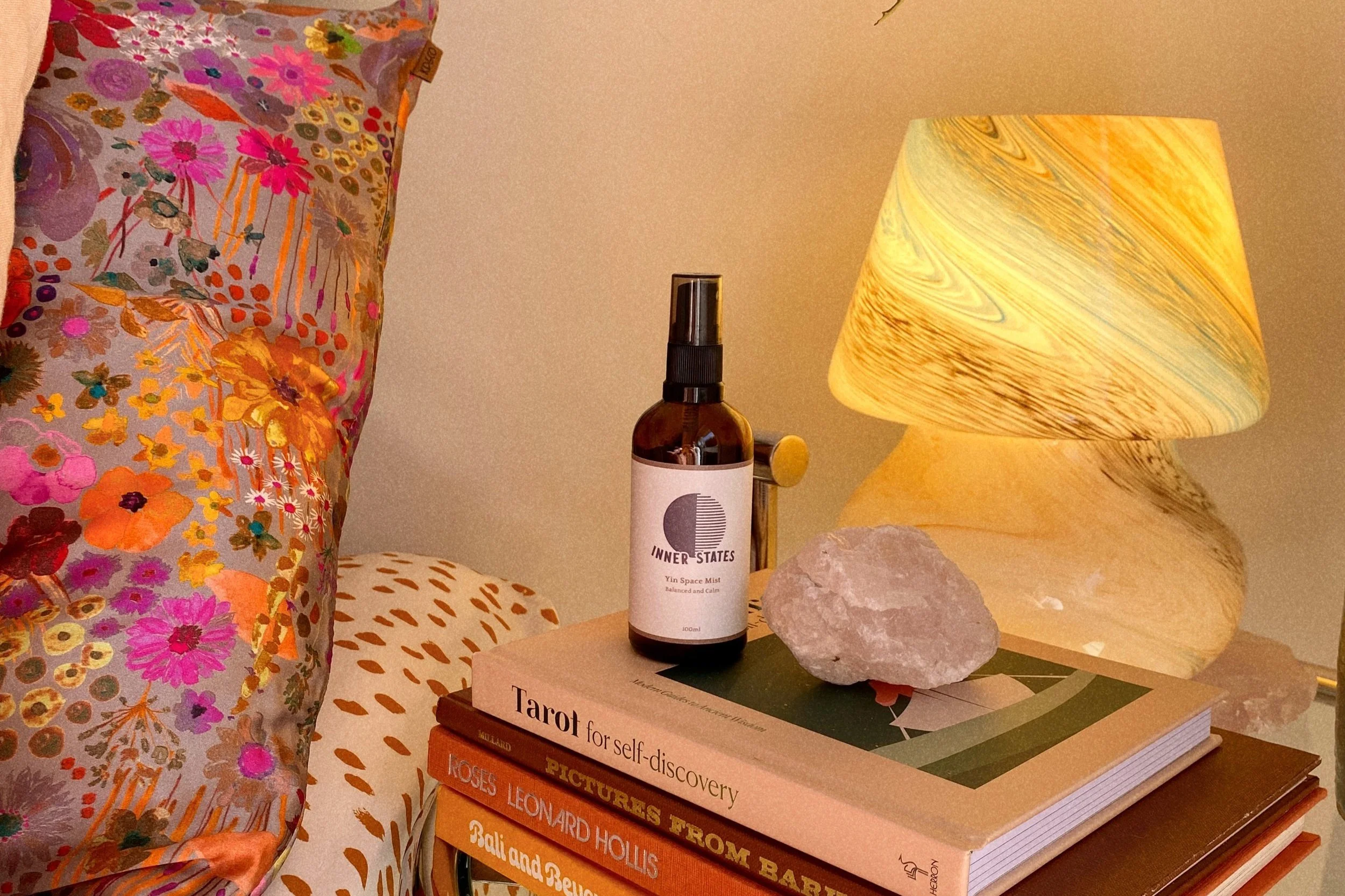 A cozy bedroom setup with a floral pillow and a stack of books on a table. A glass lamp with a marble-like pattern casts a warm glow. On top of the books, there is a spray bottle labeled 'Inner States' and a rough crystal. The scene exudes a bohemian, relaxing vibe.