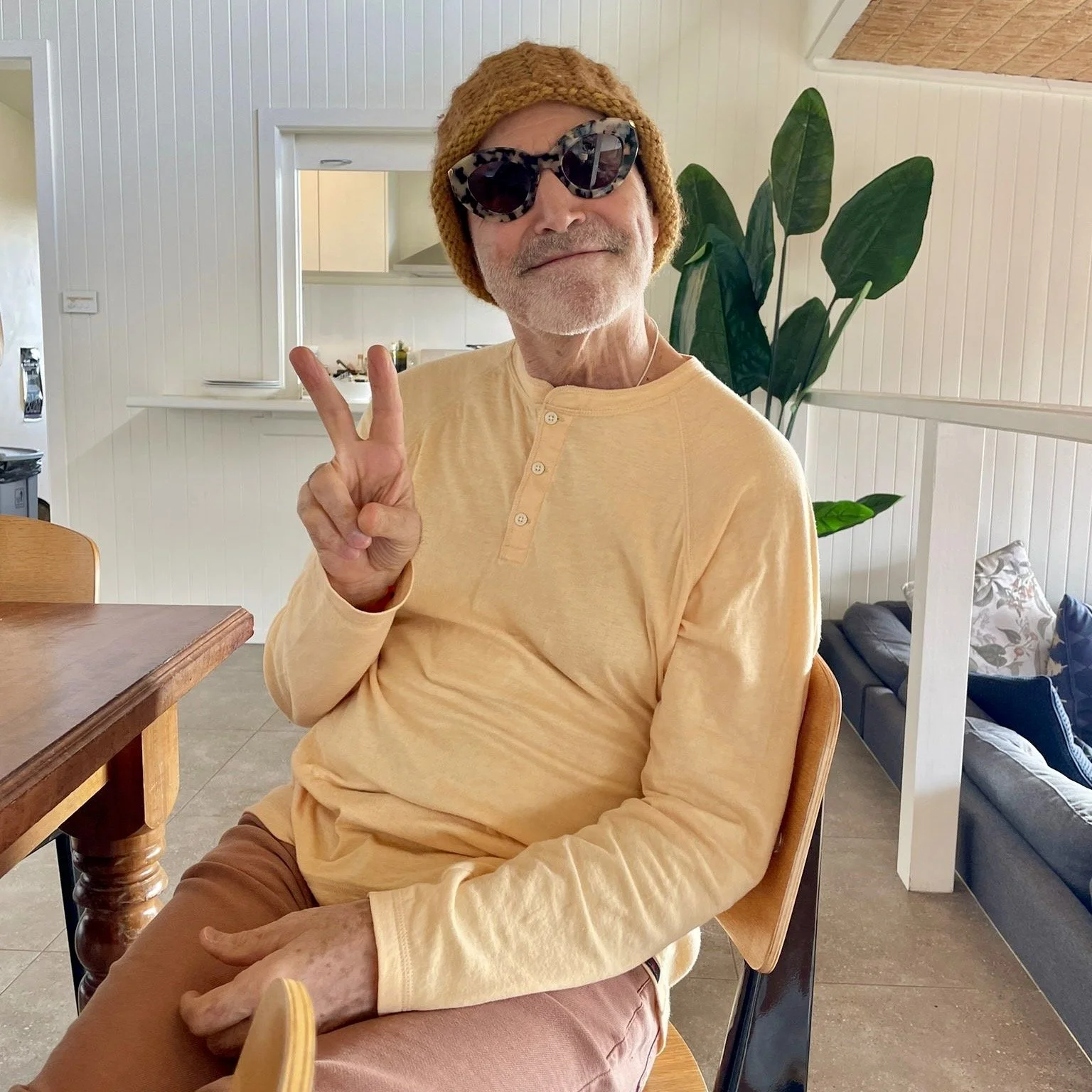Steve Ross wearing sunglasses and a beanie, making a peace sign, sitting indoors.