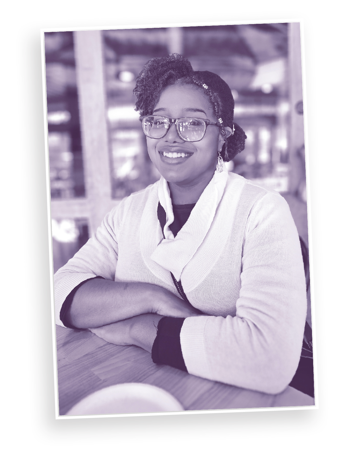 Shayla Hill (a woman with glasses) smiling, sitting at a wooden table with her arms crossed, in a casual setting with a blurred background.