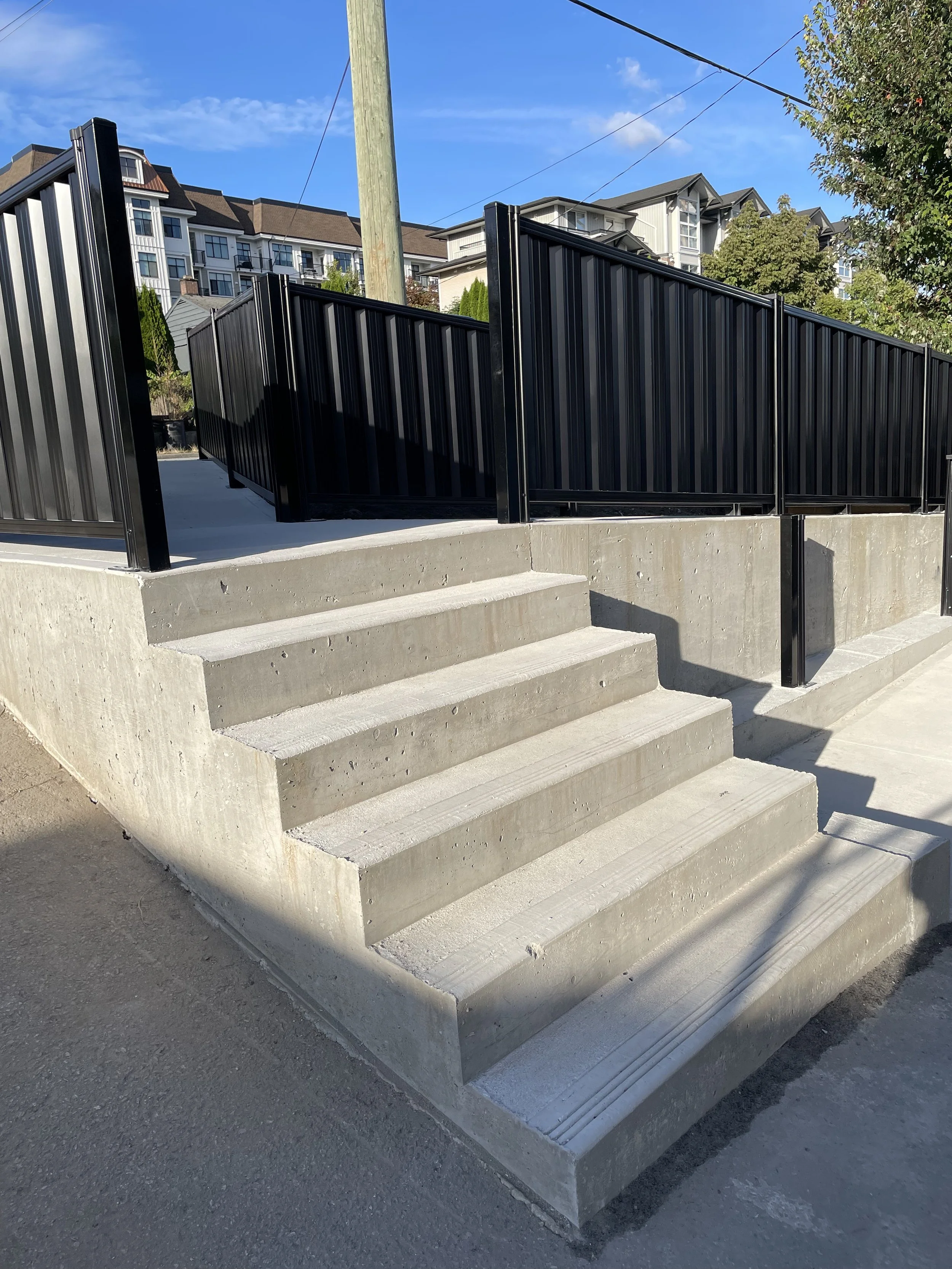 Concrete stairs leading to an elevated walkway with black metal railings, surrounded by residential buildings and a clear blue sky.