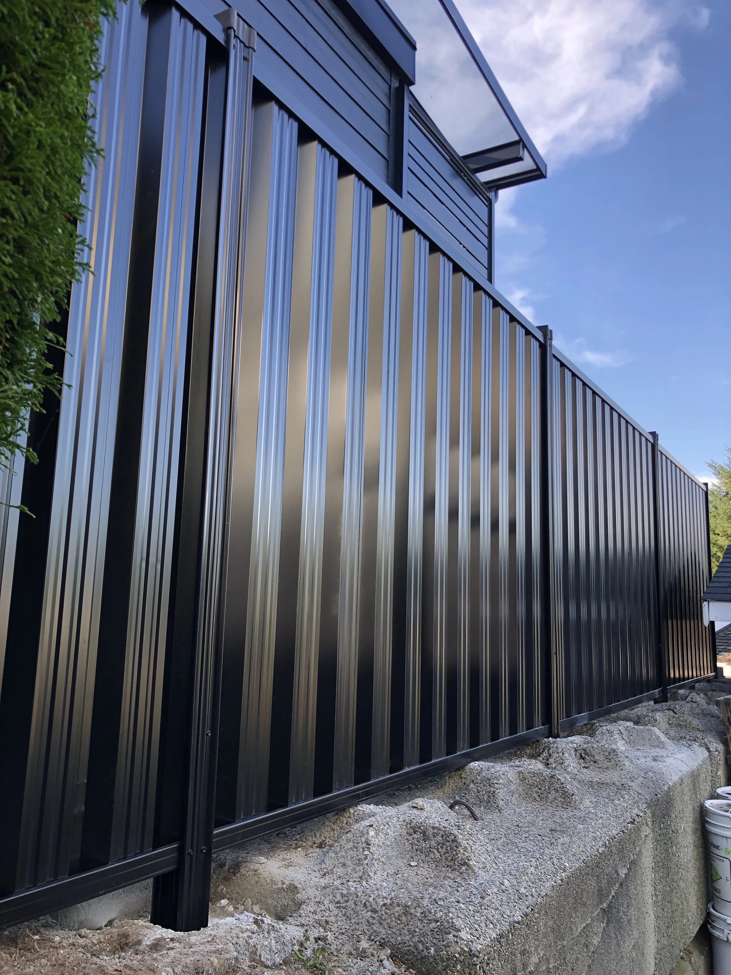 Black metal fence with vertical panels, concrete base, and adjacent green foliage under a blue sky.