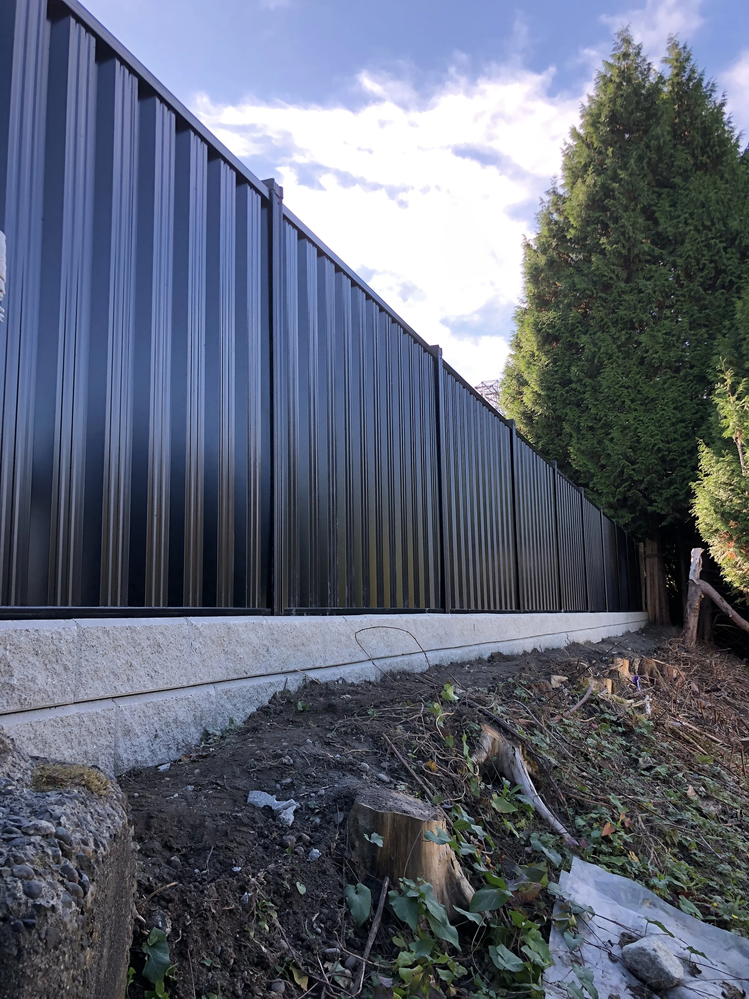 Black corrugated metal fence with concrete base next to tall evergreen trees in a landscaped yard.