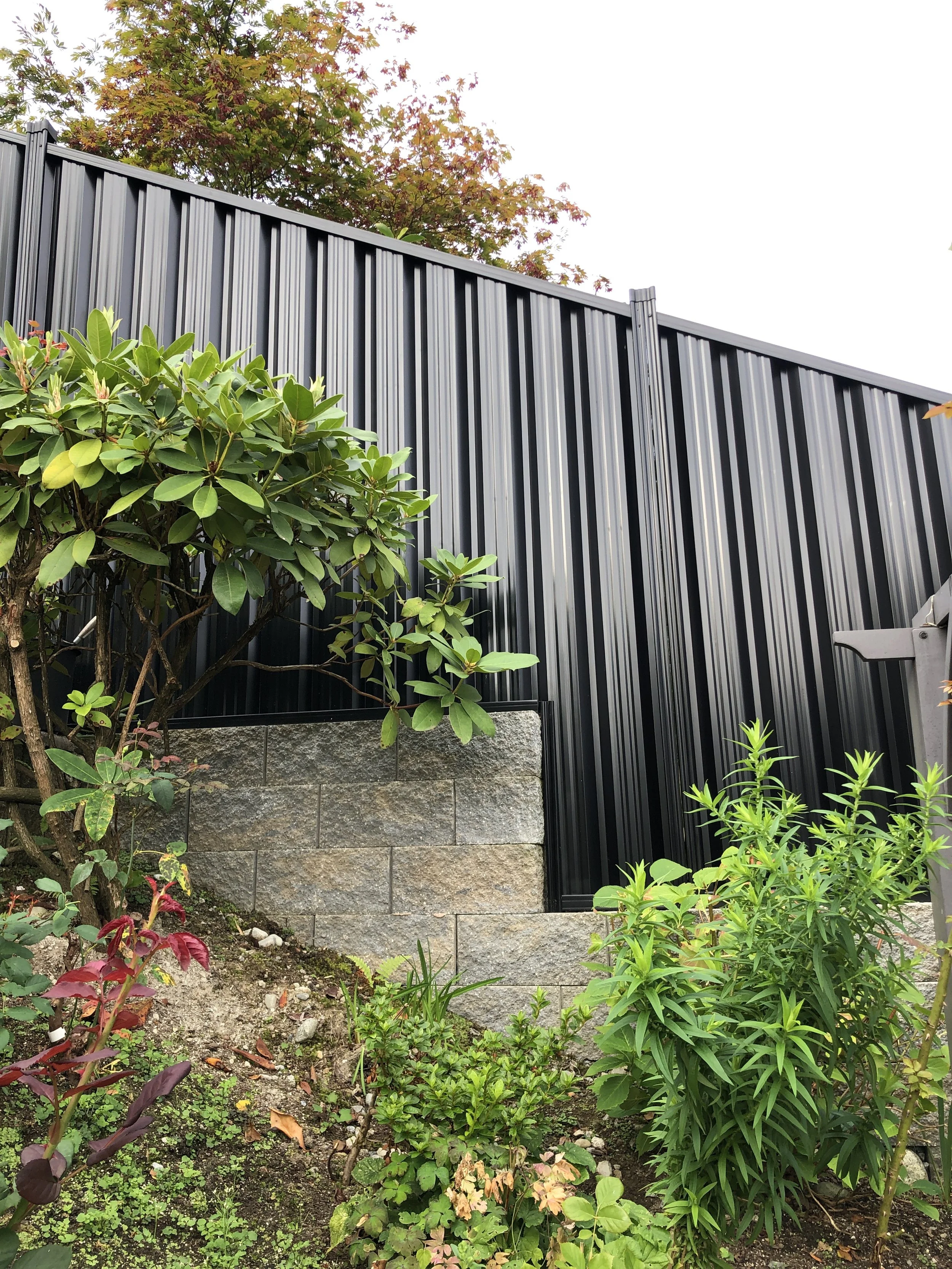 Black corrugated metal fence with a stone base and lush green foliage in the foreground, including bushes and small plants.