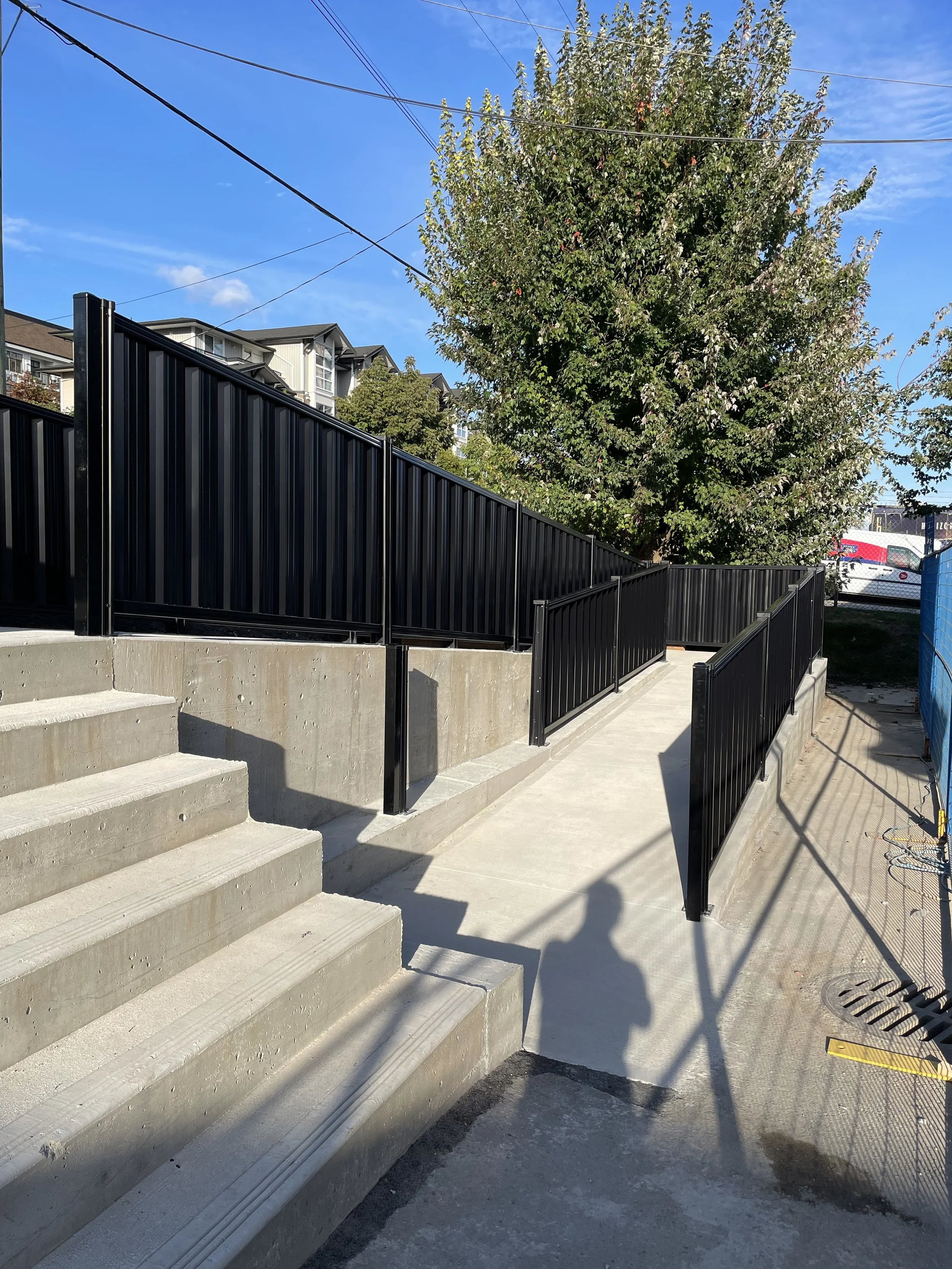 Concrete stairs and ramp with black metal railings next to a tree and buildings under a clear blue sky.