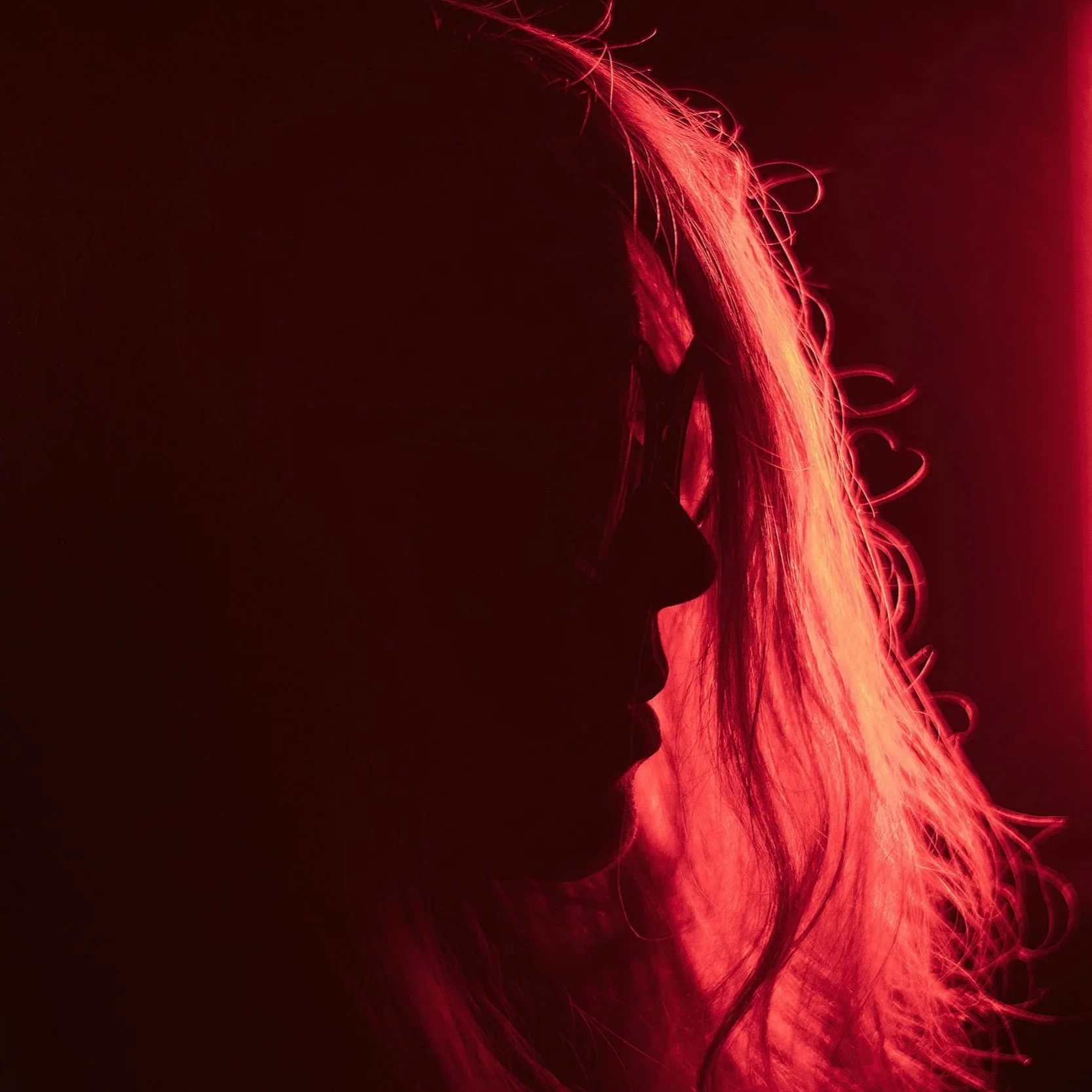 Side profile of a woman with long blonde hair illuminated in red light, with curlers in her hair and a likely makeup brush near her face.