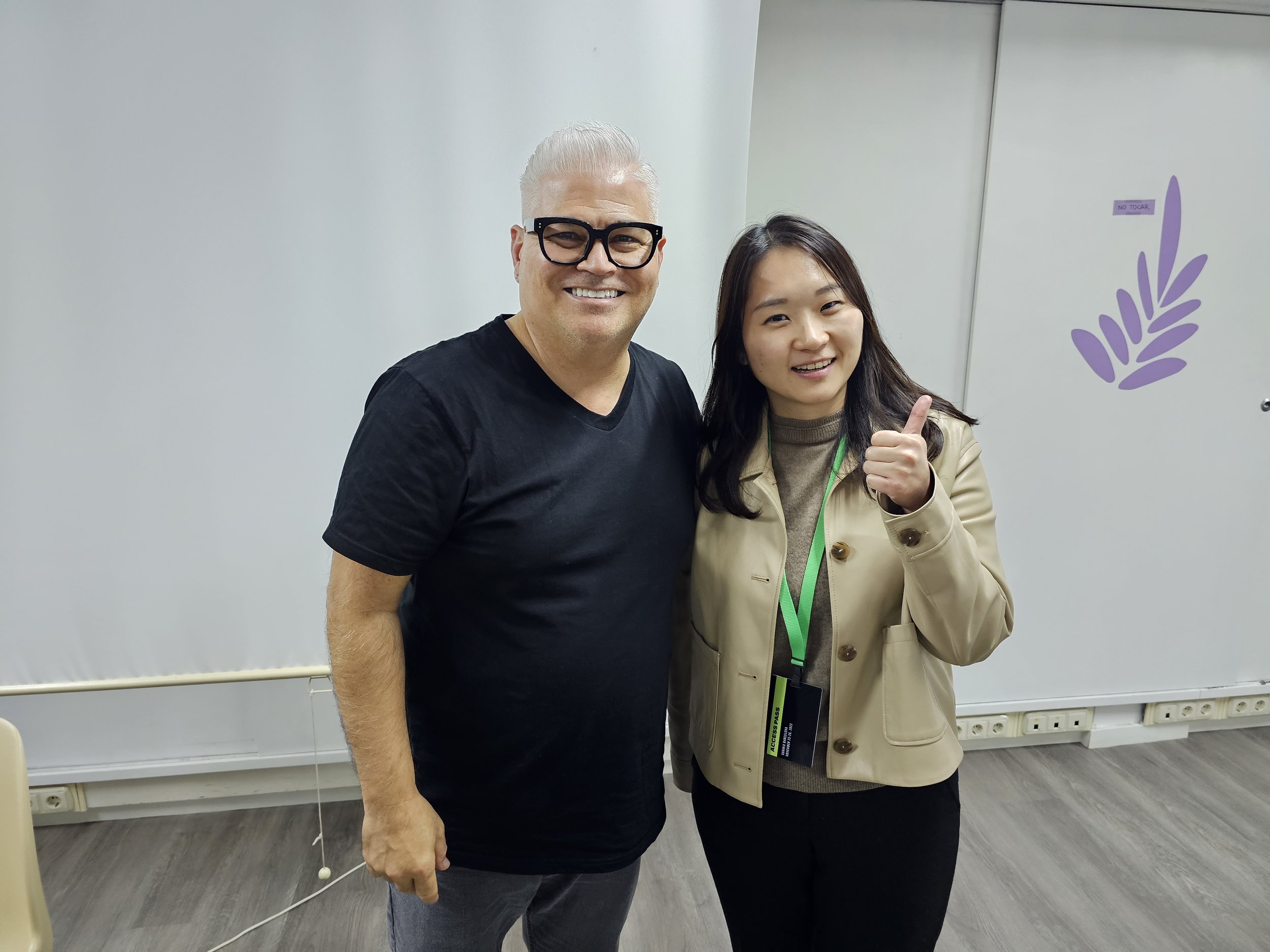 Two people standing indoors in front of a white wall with a purple leafy decorative element. One person is a man with gray hair, glasses, and a black t-shirt, smiling. The other is a woman with dark hair, wearing a beige jacket and a badge, giving a thumbs-up.