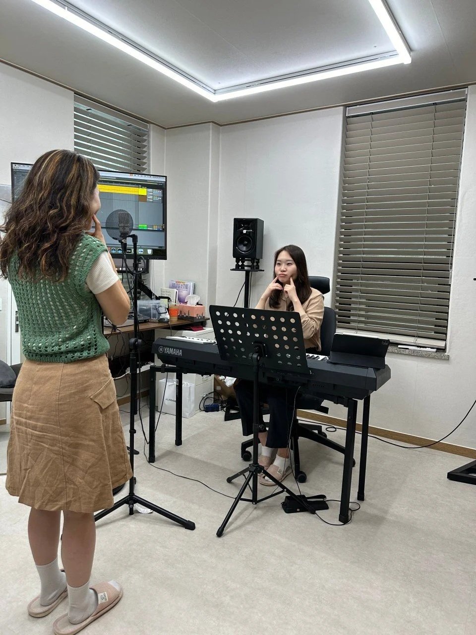 Two women in a recording studio, one standing in front of a microphone and computer monitor, the other sitting at a keyboard with music stand, in a room with window blinds and studio equipment.