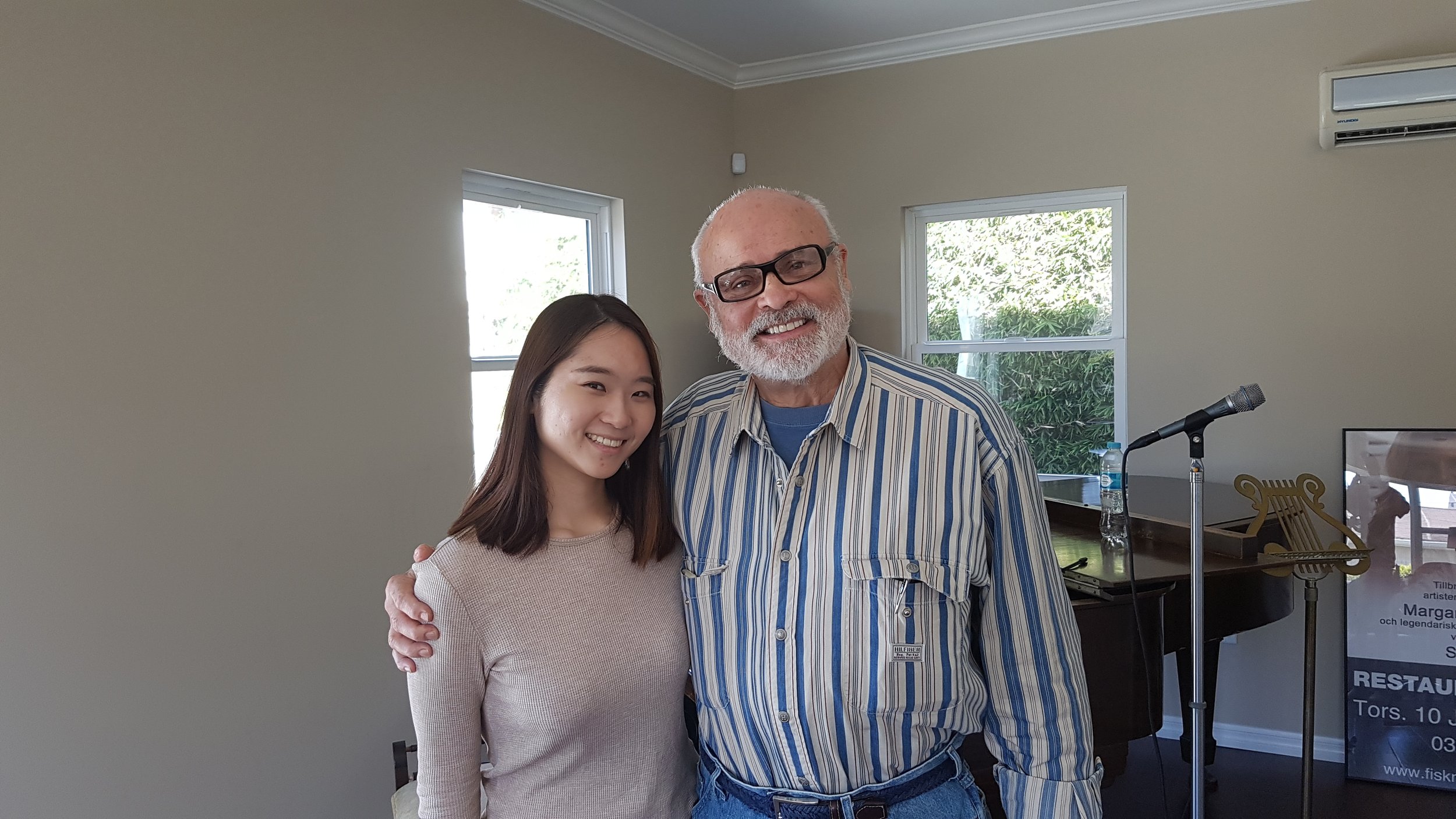 A young woman and an older man with glasses smiling and standing close together in a room with large windows, a piano, and a microphone.