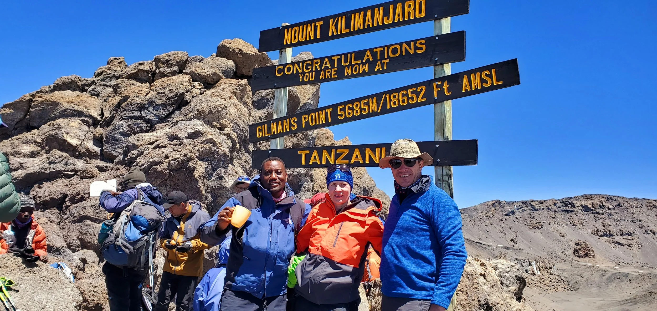 Group of hikers standing at the summit of Mount Kilimanjaro, with a large sign indicating they are at Gilman's Point, 5685 meters or 18652 feet above sea level, in Tanzania. The sky is clear and blue, and rocky terrain surrounds them.