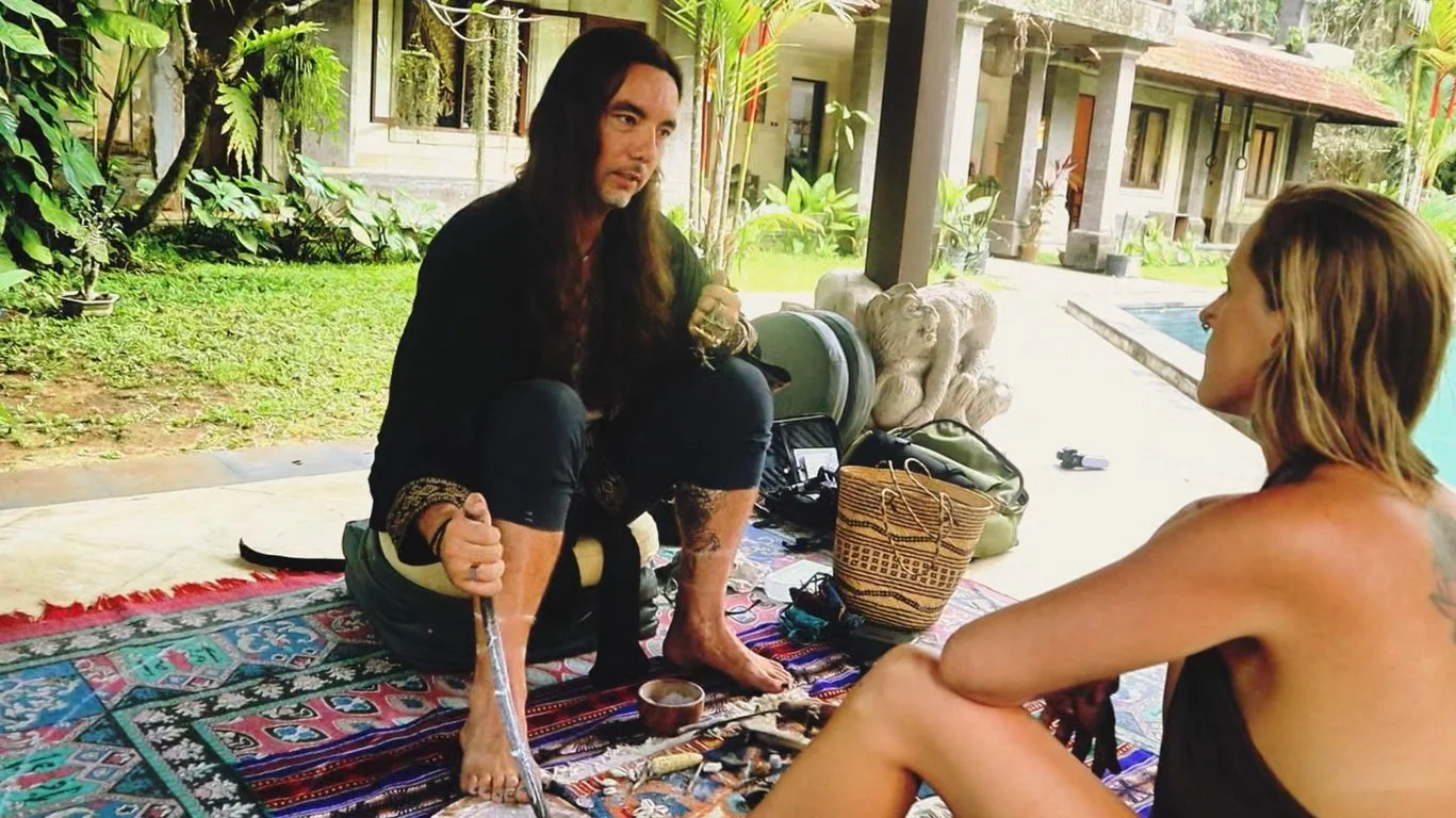 A man with long black hair sits on a woven mat holding a stick used for divination, with ritual items laid out before him. He offers a divination session to a woman with long red hair in a serene tropical courtyard with plants, stone art, and a pool.