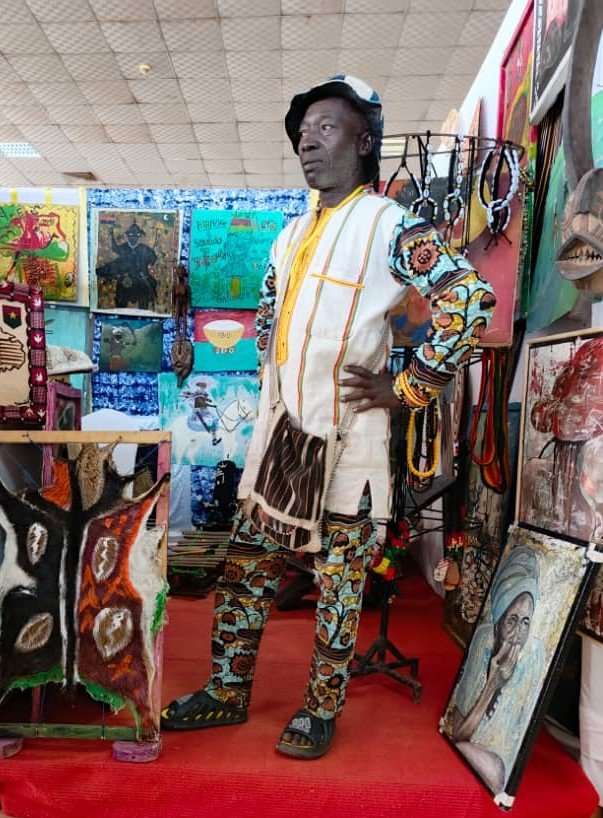 An older man dressed in colorful traditional African attire stands with hands on hips in an art display area surrounded by paintings and artwork.