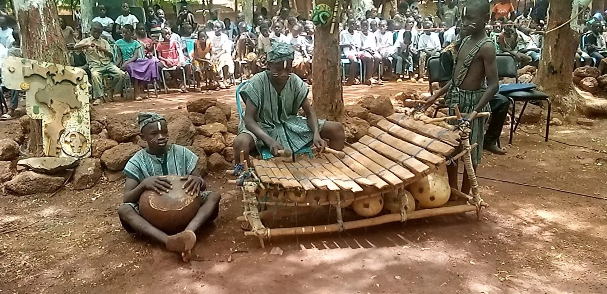 Three young men in traditional attire perform music outdoors, with one playing a drum, one playing a xylophone, and the third standing nearby. Behind them, an audience of seated people watches the performance.