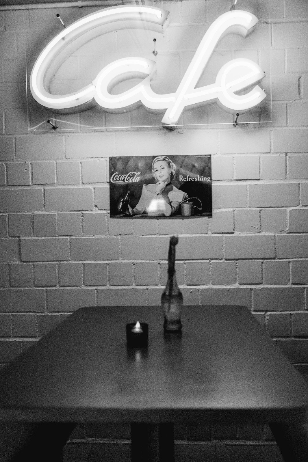Ein schwarz-weiß Bild zeigt eine Tafel mit einem Neonlicht, das das Wort 'Café' leuchtet, eine Wand mit einer alten Coca-Cola Werbung, ein Tisch mit einer Vase und einer kleinen Kerze in einem dunklen Raum.