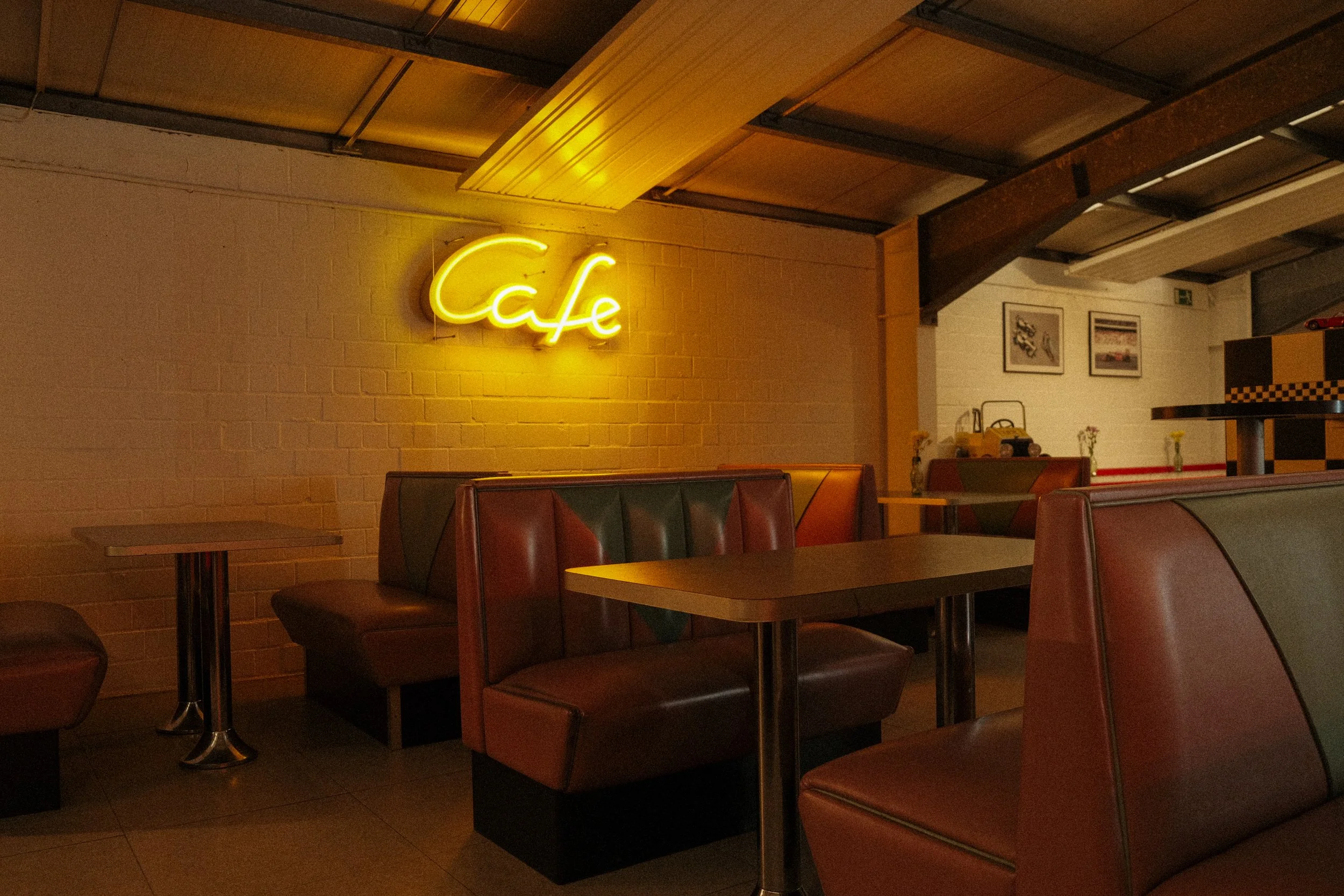 Innenansicht eines Cafés mit braunen Ledersofas, Tischen und einer gelben Neon-Schrift, die 'Café' an eine Wand leuchtet.