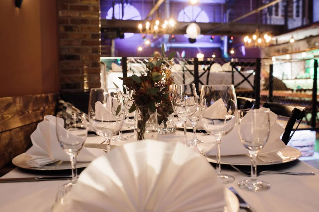 Ein festlich gedeckter Tisch mit weißen Napkins, Weingläsern und einer Blumenvase in einem Restaurant mit Lichterdekoration an der Decke.