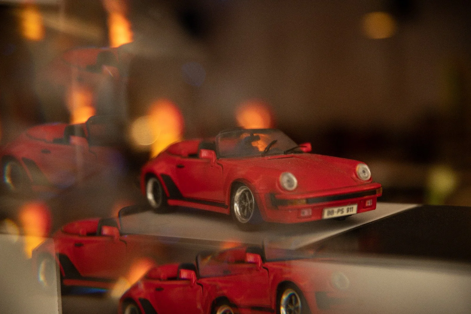 Ein rotes Modellauto eines Porsche 911 auf einem reflektierenden Oberflächen. Im Hintergrund sind bunte Lichter und weitere reflektierende Flächen sichtbar.