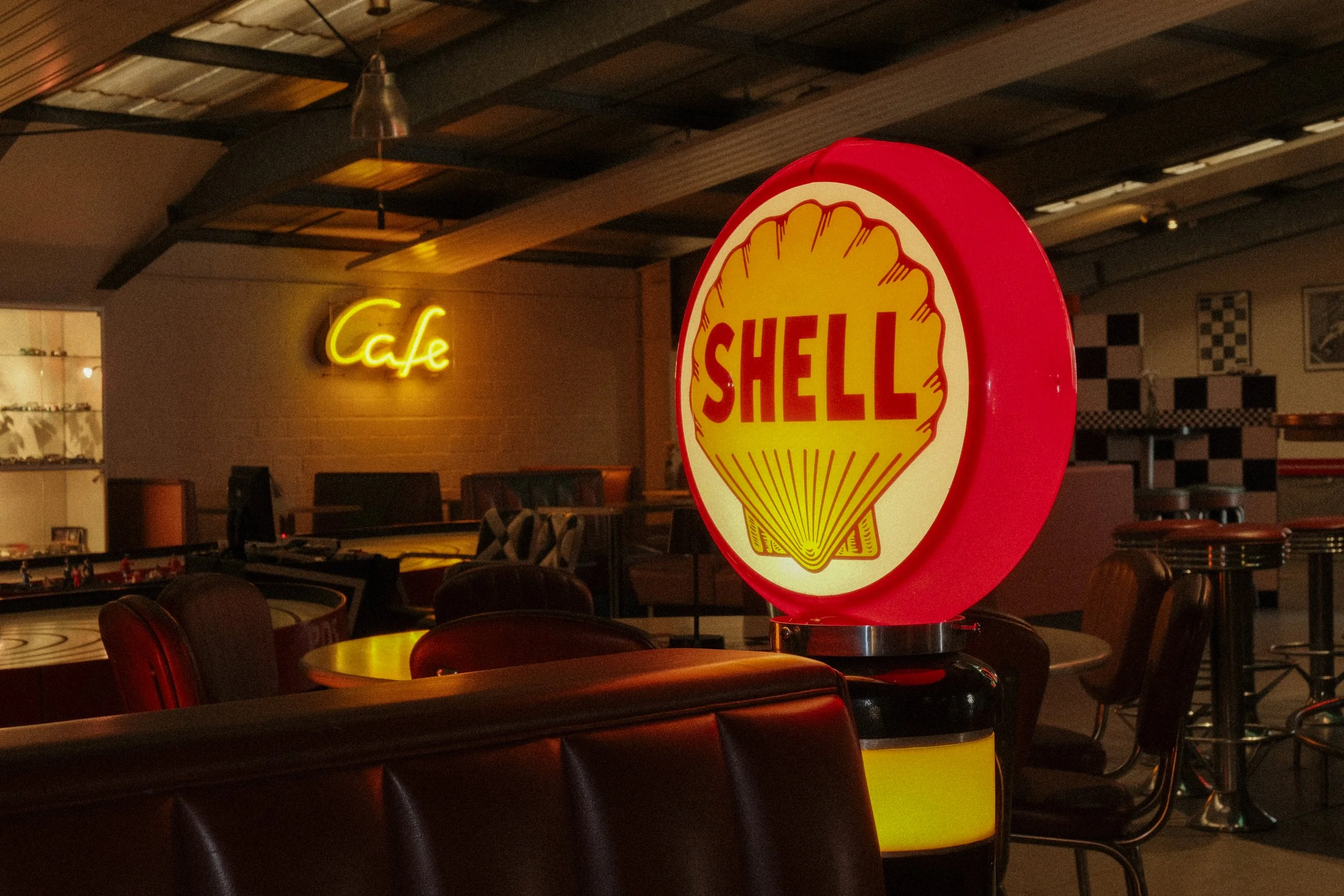 Ein neonbeleuchtetes Café mit einem Shell-Logo, das an einer Bar platziert ist, und einem gelben "Café"-Schild an der Wand im Hintergrund.