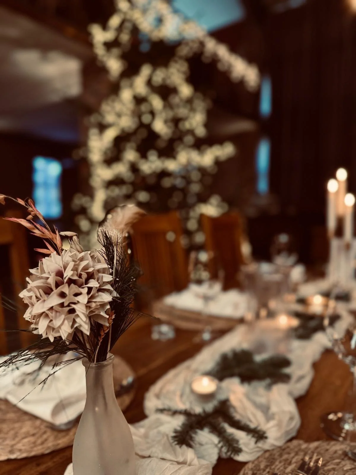 Ein festlich dekorierter Tisch mit Blumen und Kerzen, im Hintergrund ein Weihnachtsbaum mit weißen Kerzen in einer Holzhütte.