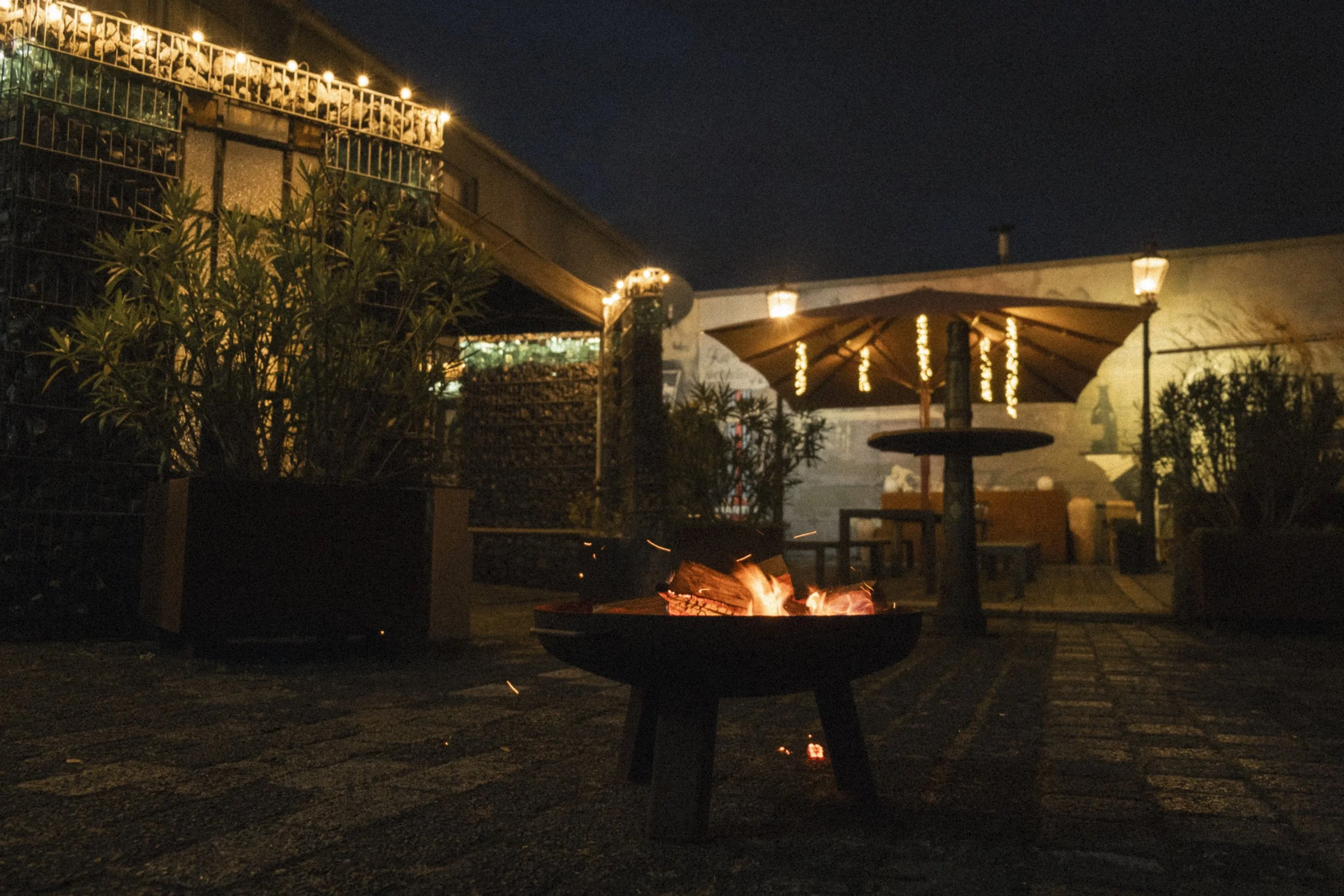 Gemütlicher Außenbereich bei Nacht mit Feuerstelle, Sitzgelegenheiten, Sonnenschirm und Lichterketten.