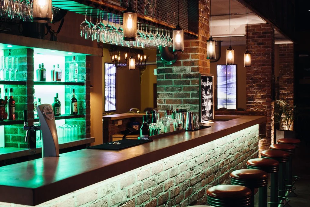Ein Barbereich mit einem Brick-Design, beleuchtet mit warmen und grünen Lichtern, und Barhockern am Tresen.