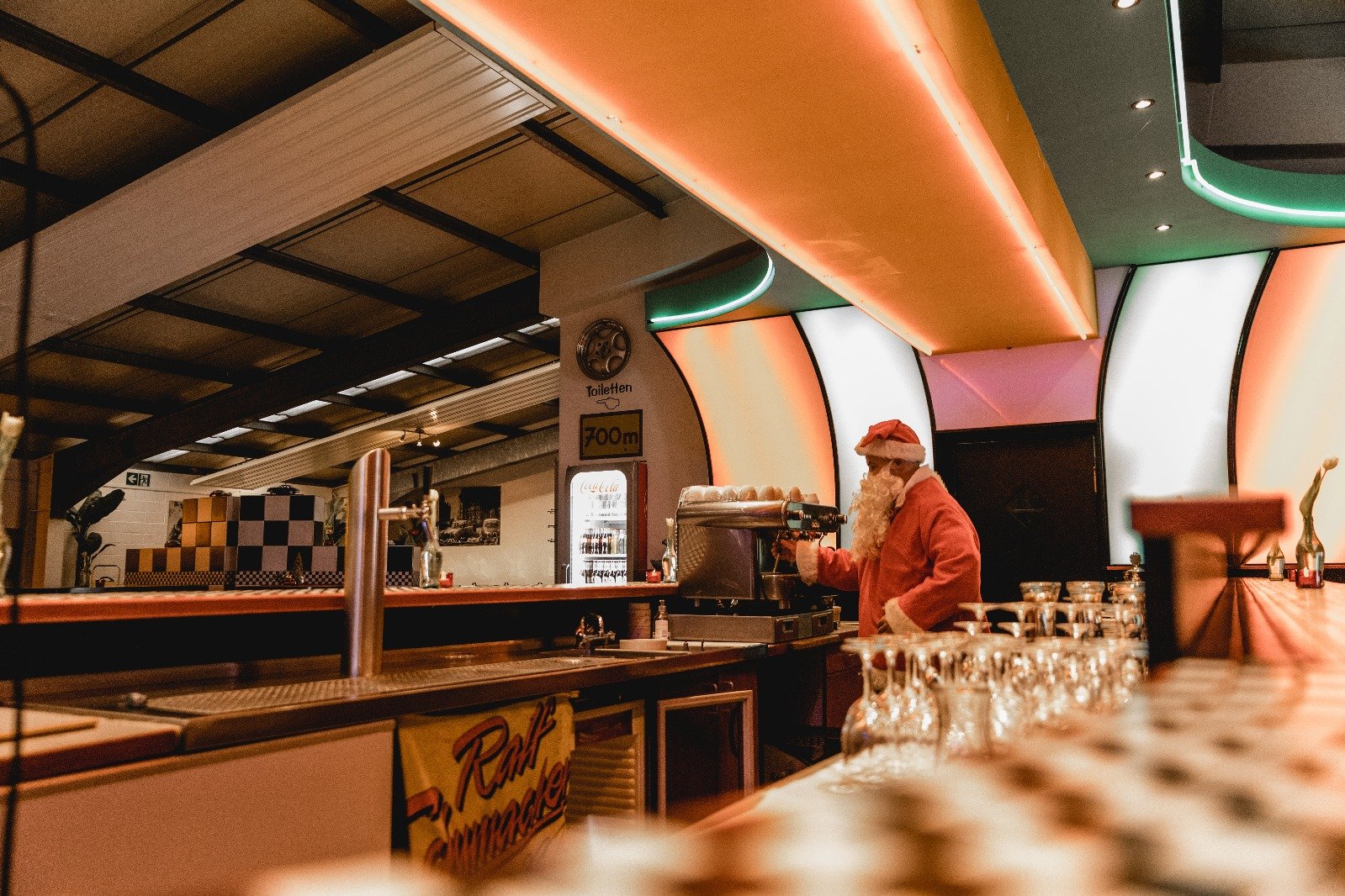 Ein Mann in einem roten Santa-Kostüm steht hinter einer Bar und serviert Getränke. Die Bar ist in einem modernen Restaurant oder Club mit leuchtenden, bunten Lichtdesigns im Hintergrund.