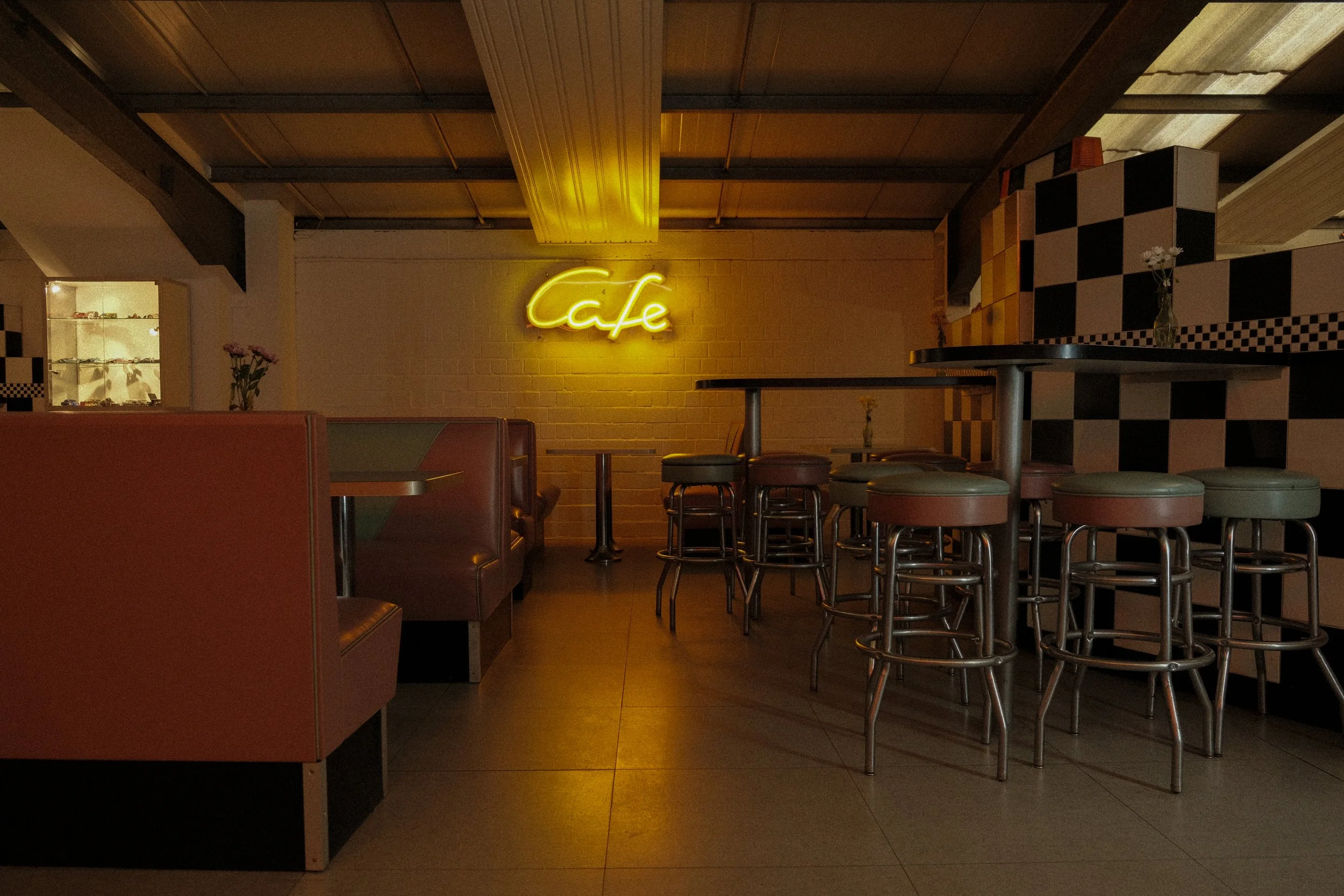 Innenraum eines Cafés mit hoher Decke, neonfarbenes Biergst â€žcafeâ€ an der Wand, Barhocker um einen hohen Tisch, gepolsterte Sitzbänke, Vasen mit Blumen, schwarz-weiß gemusterte Wände.