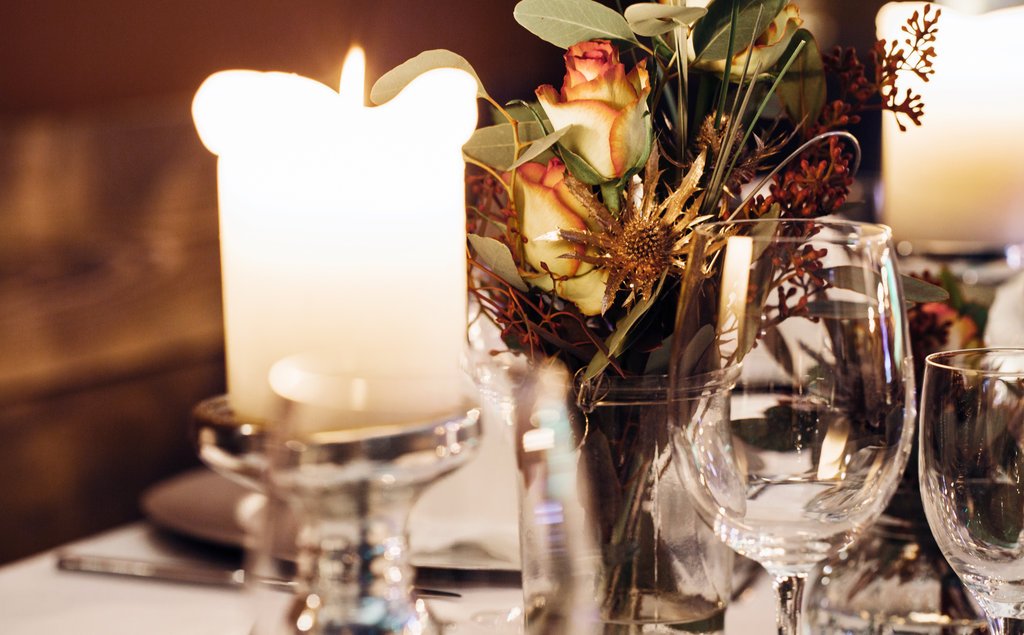 Tischdeko mit Kerze, Blumenstrauß in Vase, Weingläser, Teller, Besteck auf weißem Tischtuch