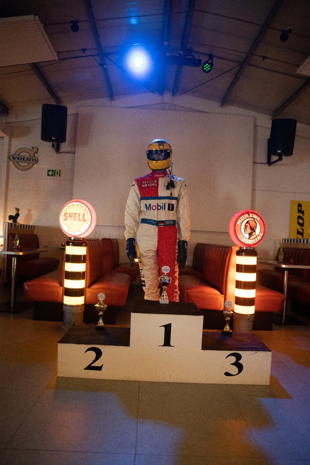 Souveräner Formel 1 Sieger auf dem ersten Platzpodest, umgeben von Trophäen, mit einem Rennfahrer in Rennmontur und Helm, im Hintergrund Markenlogos wie Shell und Mohawk Gasoline.