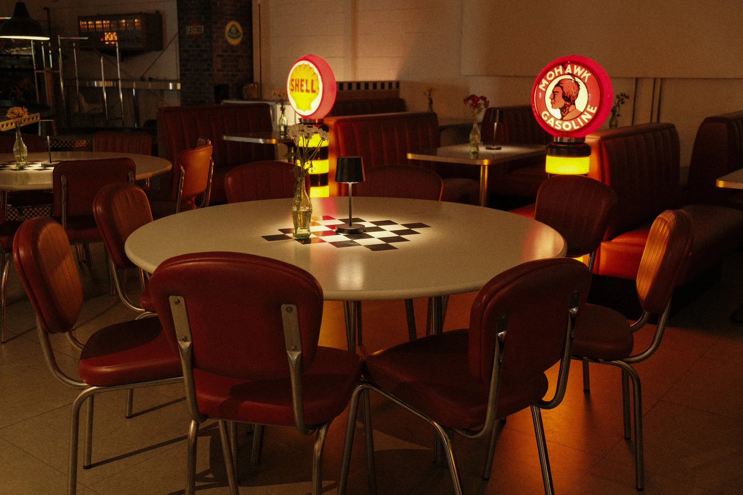 Ein gemütliches Lokal mit mehreren Tischen, Rotstühlen und Vintage-Illuminationslichtern im Hintergrund.