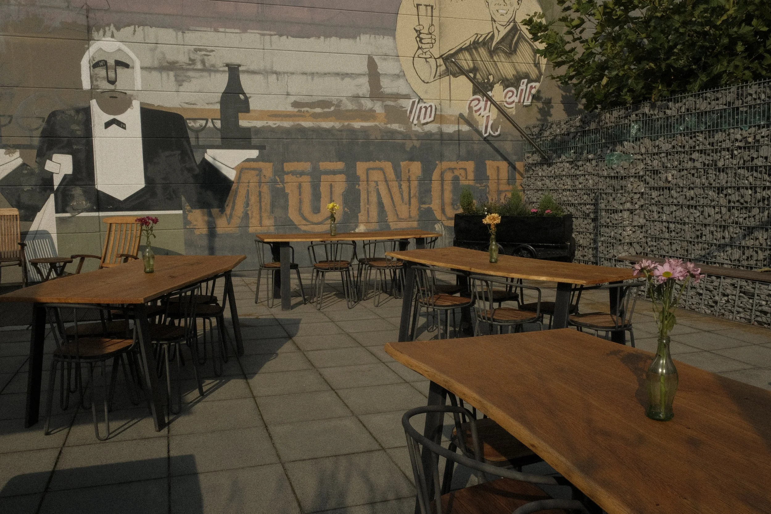 Blick auf einen Außenbereich eines Restaurants mit Holztischen, glasvasen mit Blumen, Wandbild mit einem männlichen Servicepersonal, das ein Glas hält, und der Schriftzug 'Münch' im Hintergrund.
