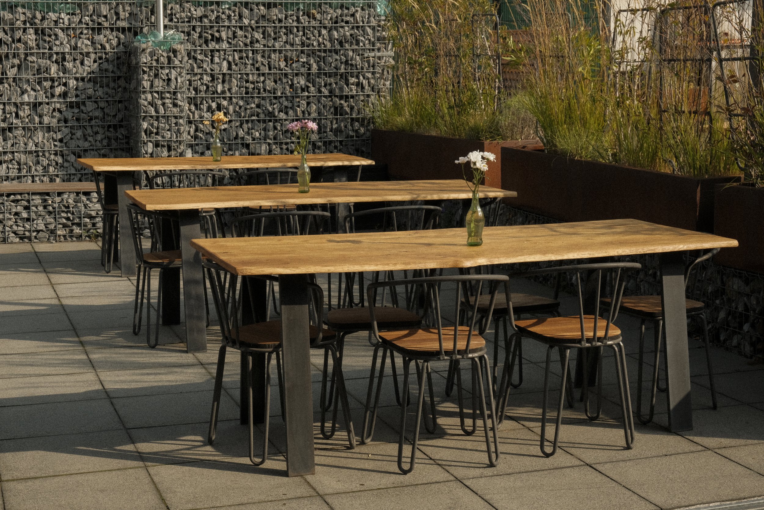 Drei Holztische mit jeweils einem Blumenvasen, umgeben von schwarzen Stühlen auf einer Terrasse mit Fliesenboden, im Hintergrund eine Metallwand und bepflanzte Begrenzung.