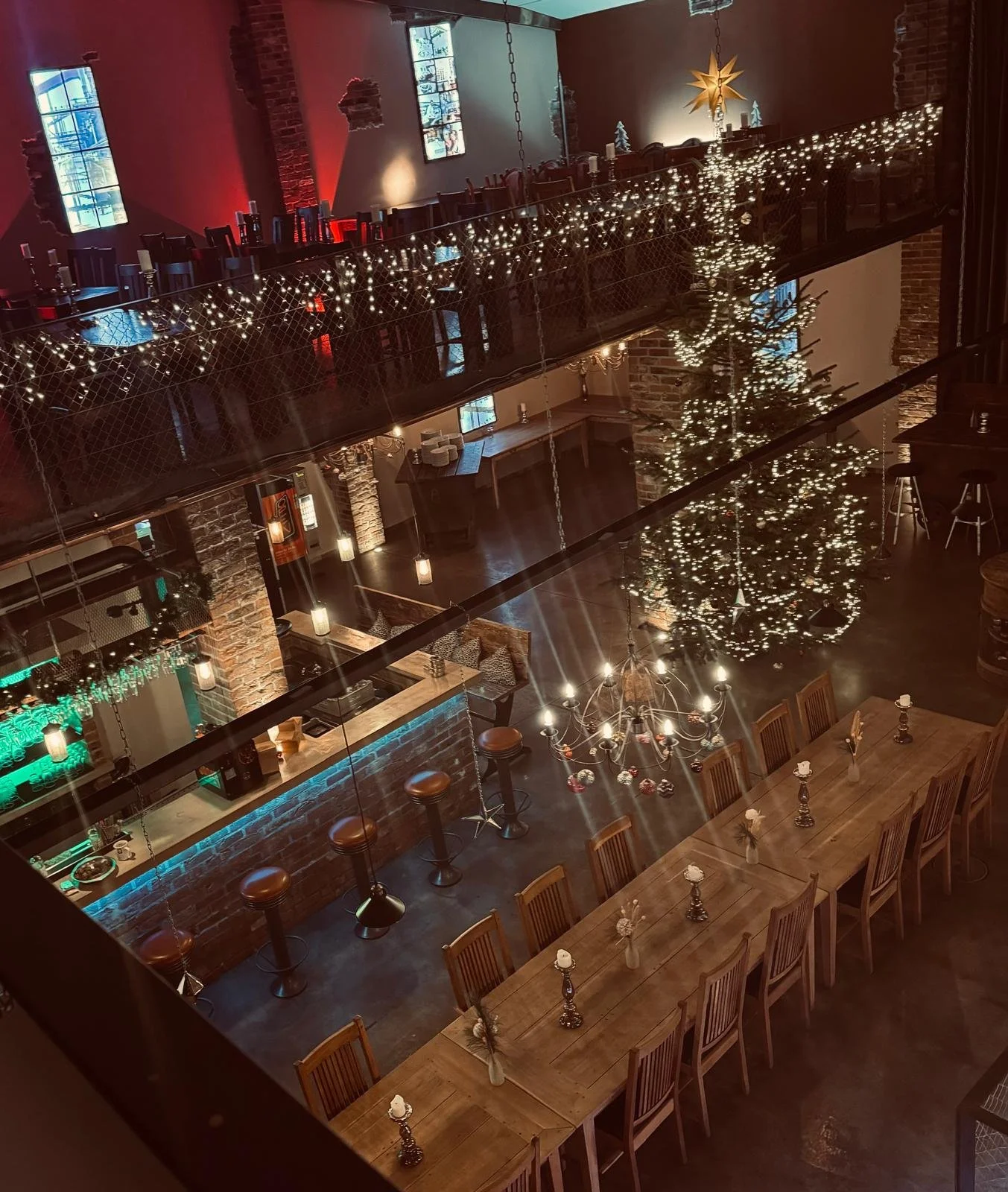 Ein festlich dekorierter Innenraum eines Restaurants mit Weihnachtsbaum, Lichterketten, einem Kronleuchter und einem langen Holzdiamond-Tisch mit Kerzen. Oben befindet sich eine zweite Ebene mit Tischen, ebenfalls mit Weihnachtsdekoration, und große 