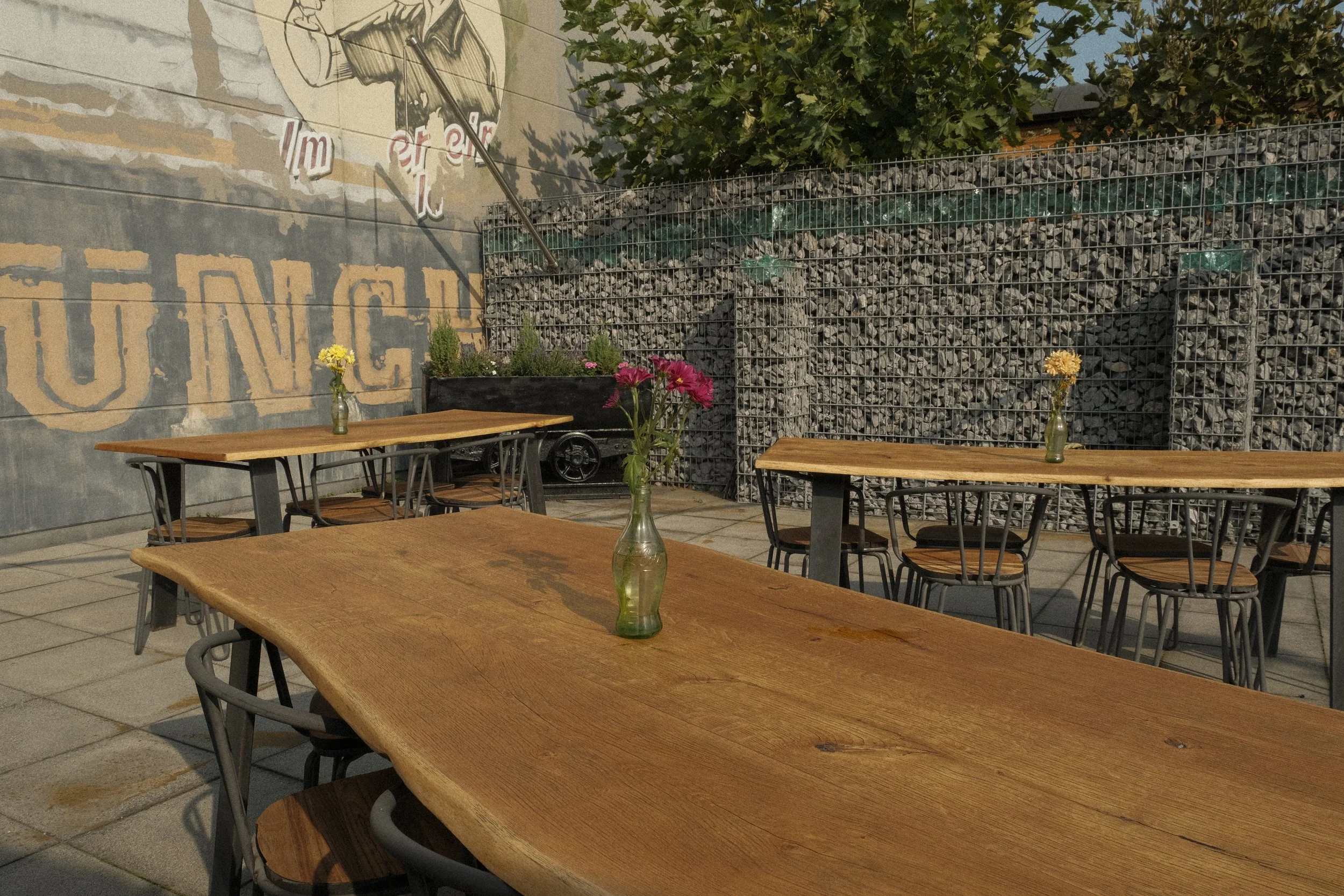 Ein Außenbereich mit Holztischen und Stühlen, auf jedem Tisch steht eine kleine Vase mit bunten Blumen. Im Hintergrund eine Mauer mit Graffiti und eine Gabionenwand.