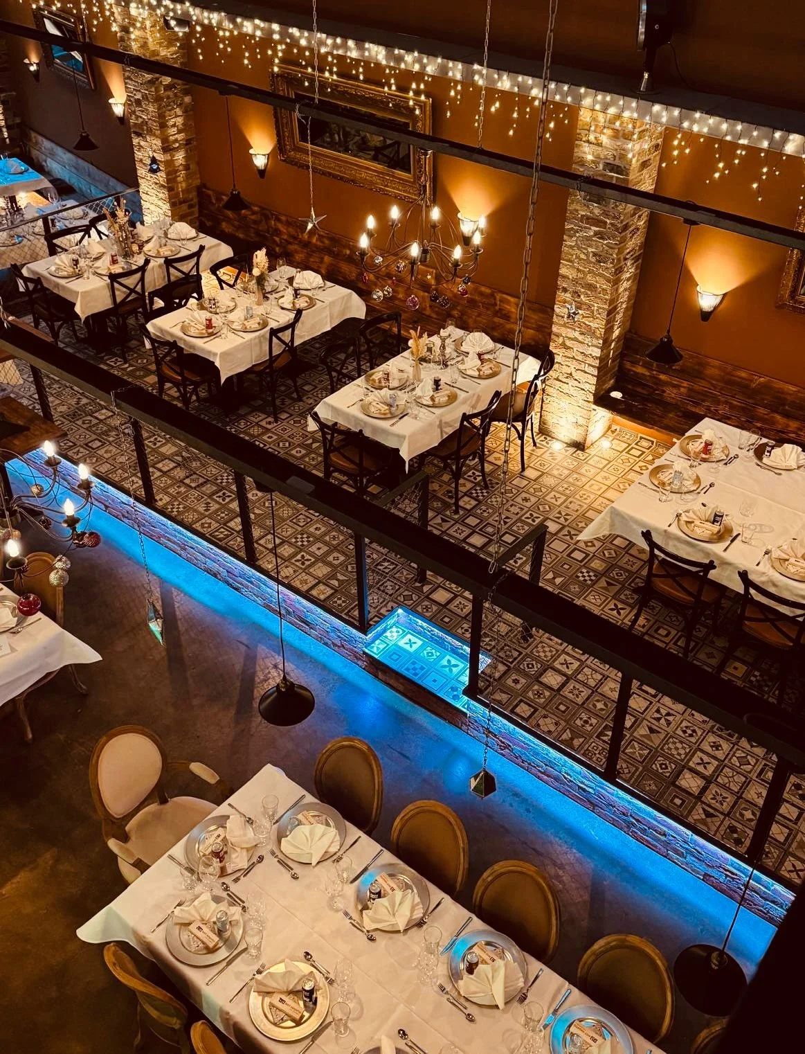 Gut gedeckter Esstisch im Restaurant mit weißen Tischdecken, Besteck, Gläsern, weißen Stoffservietten, kleinen Tischkarten, zuvorkommender Beleuchtung, und einer zweiten Ebene mit weiteren Tischen, gepflasterten Wänden und warmem Licht.