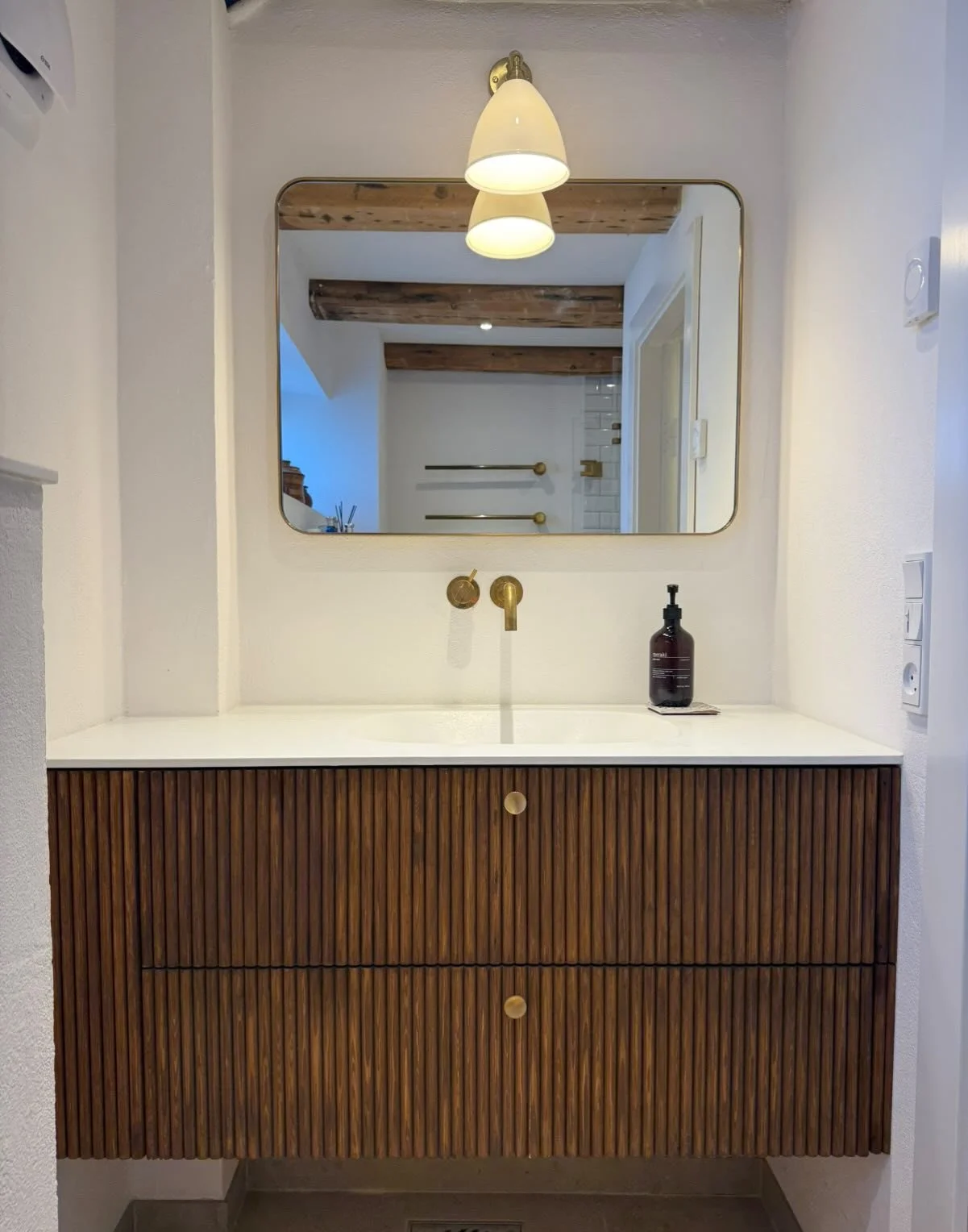 Badeværelse med hvid væg, træskab med lodrette striber, hvid vask, spejl, væglampe, håndsæbe dispenser, og rør til vand.