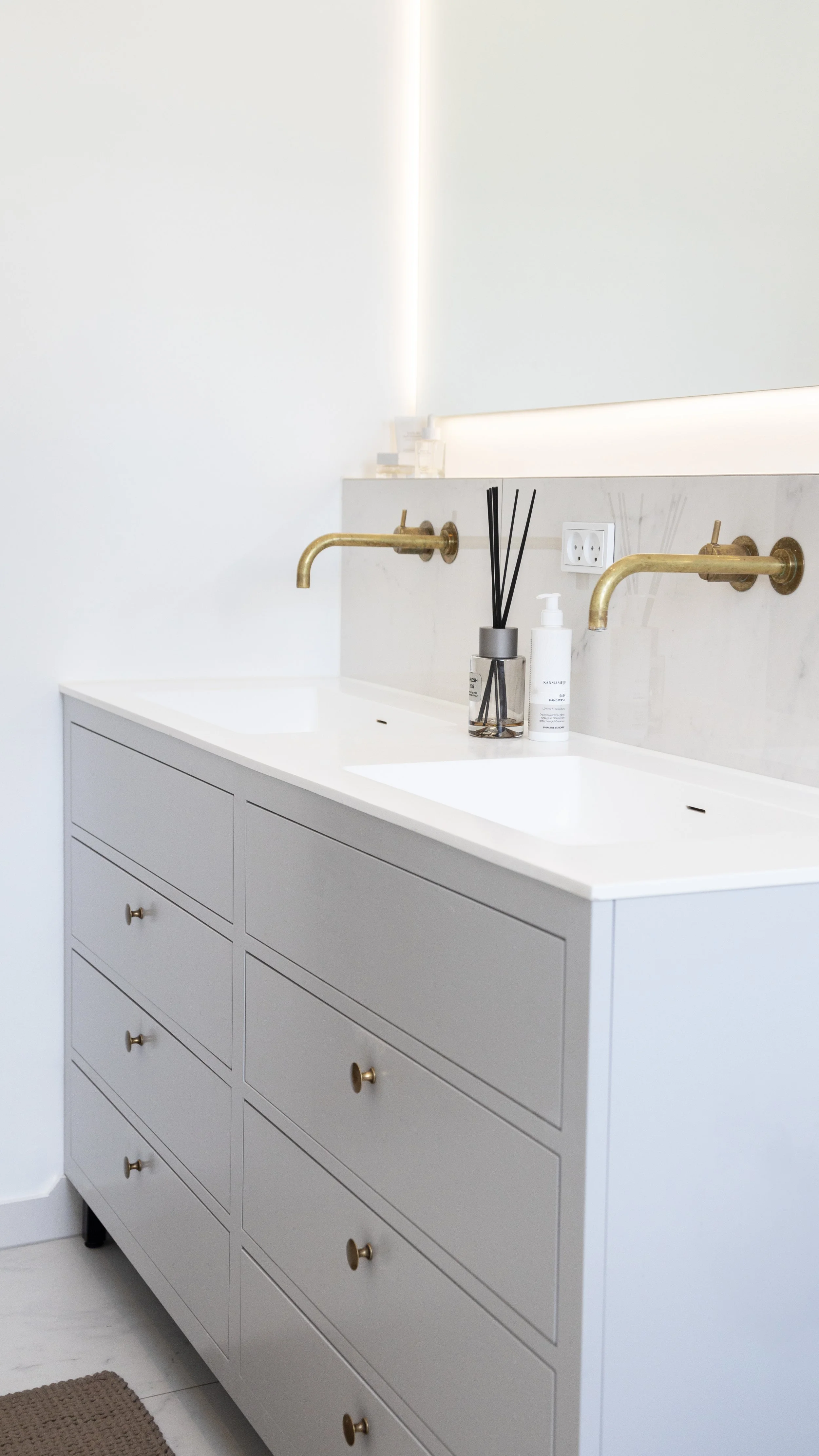 Badeværelse med hvid skabsdobbeltvask, to messingarmaturer, et spejl med baggrundsbelysning, en dispenser med håndsæbe, en glas beholder med duftpinde, stikkontakt på væggen.